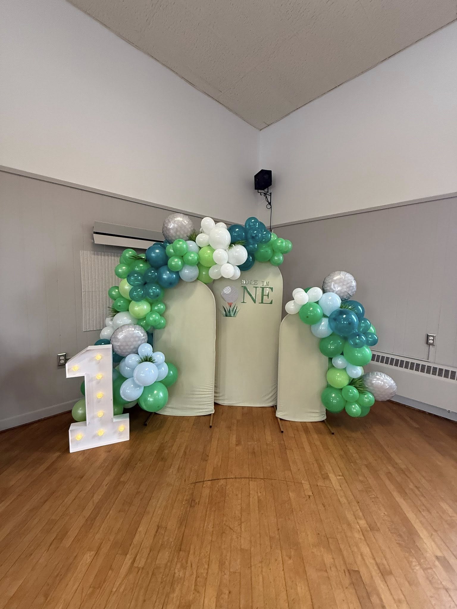 Golf-themed first birthday party backdrop with green and white balloons, arch panels, and a number one marquee light.