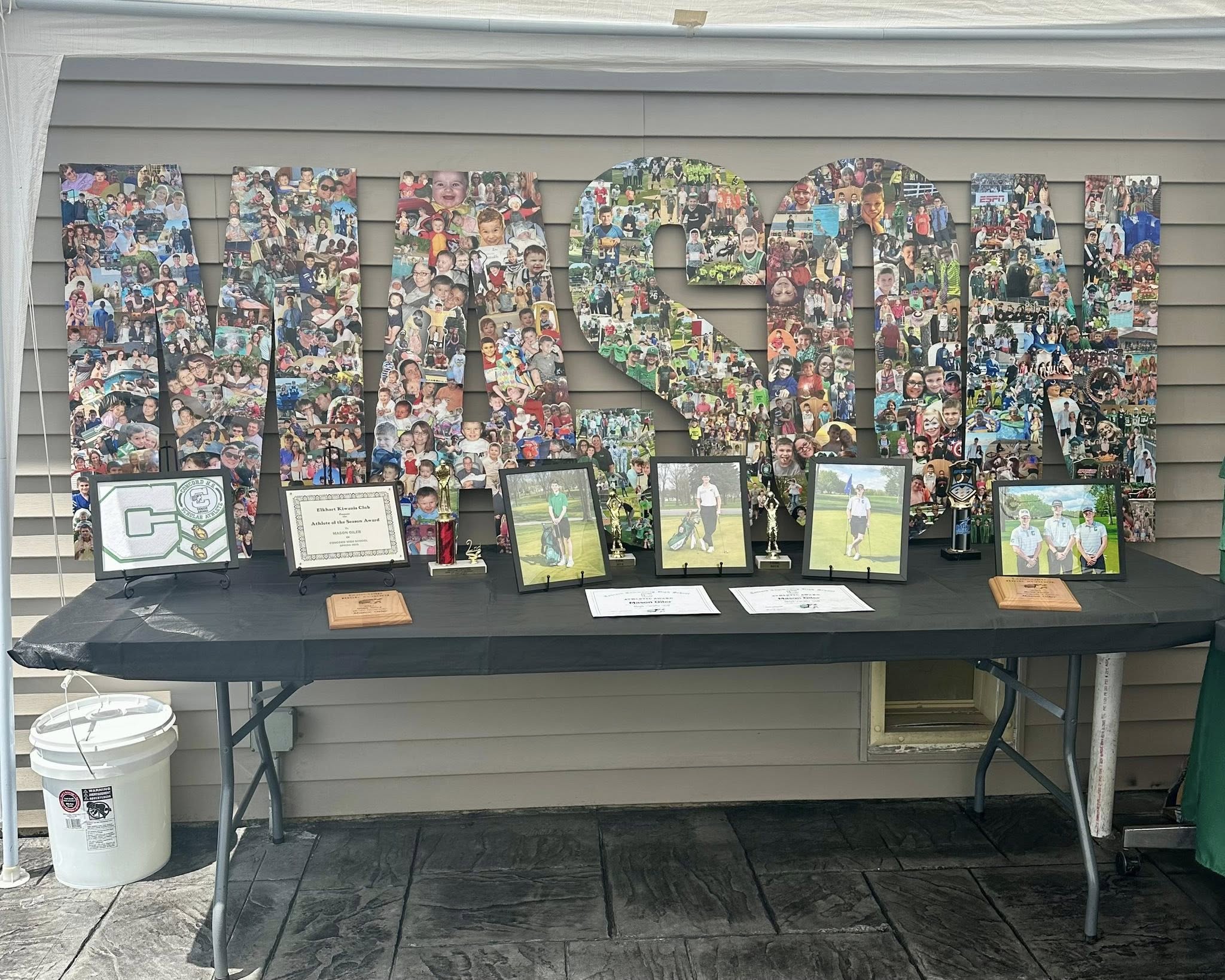 A party display featuring the name MASON spelled out in large photo collage letters with athletic awards, trophies, and portraits on a black table.