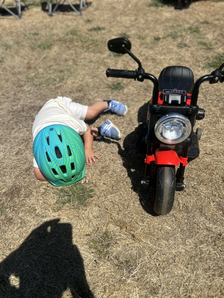 Toddler wearing a bright green helmet lying facedown on dry grass next to a red toy motorcycle.