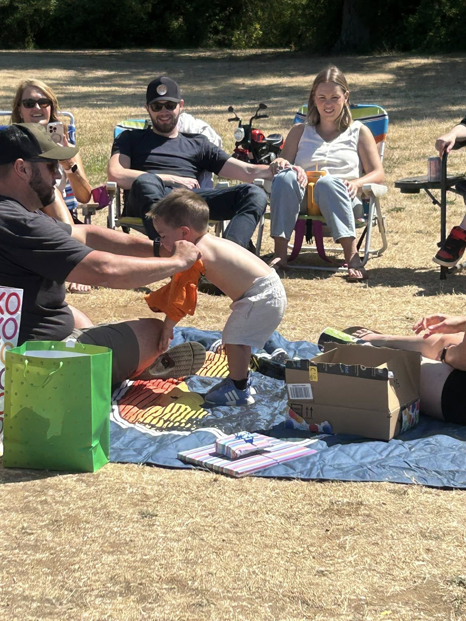 A man helping a young toddler put on an orange t-shirt outdoors while family sits in lawn chairs.