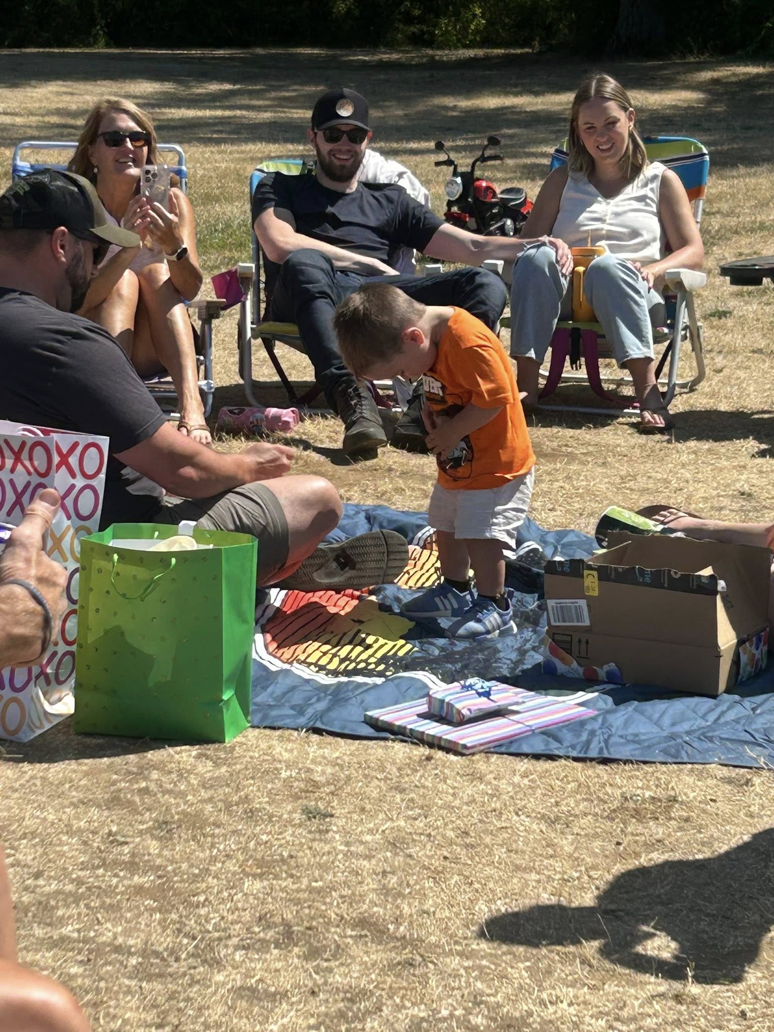 A small child in an orange t-shirt standing on a picnic blanket surrounded by family and birthday gifts in a park.
