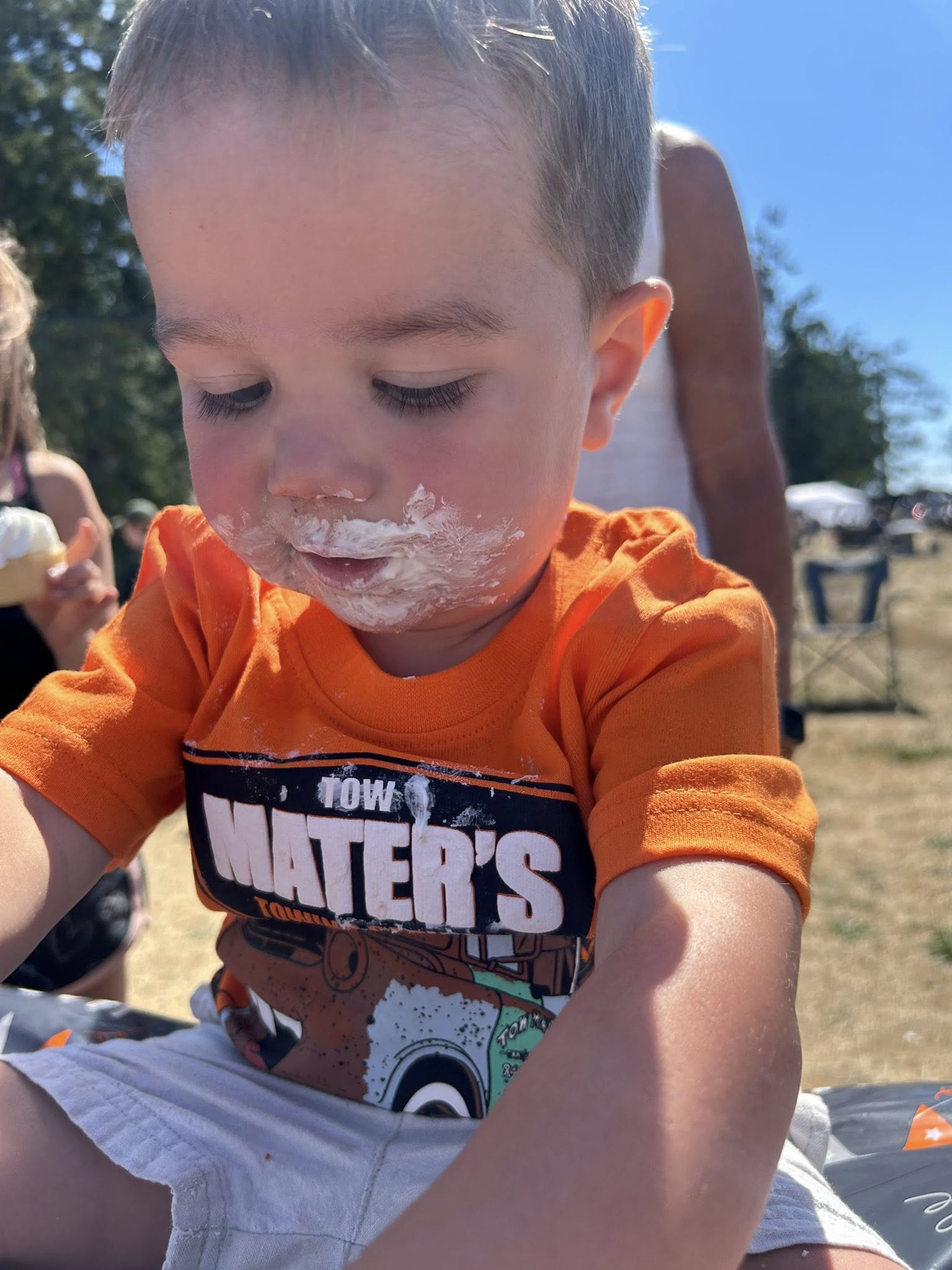Toddler wearing a Cars Mater t-shirt with ice cream on his face while eating outside.