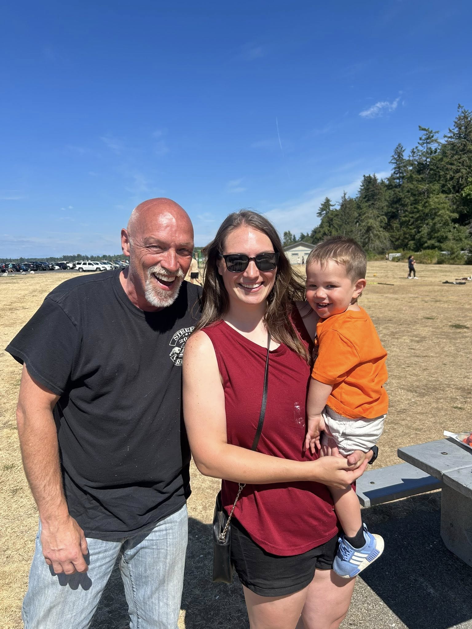A smiling man, woman, and toddler standing together at an outdoor park on a sunny day.