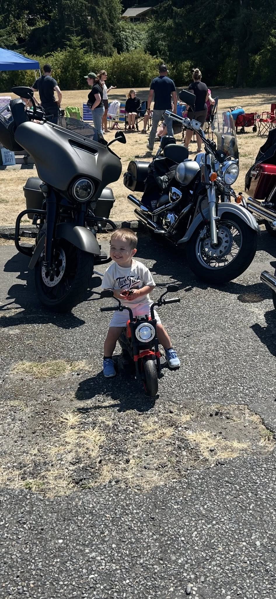 A smiling young boy sitting on a red toy motorcycle in front of full-sized motorcycles at an outdoor event.