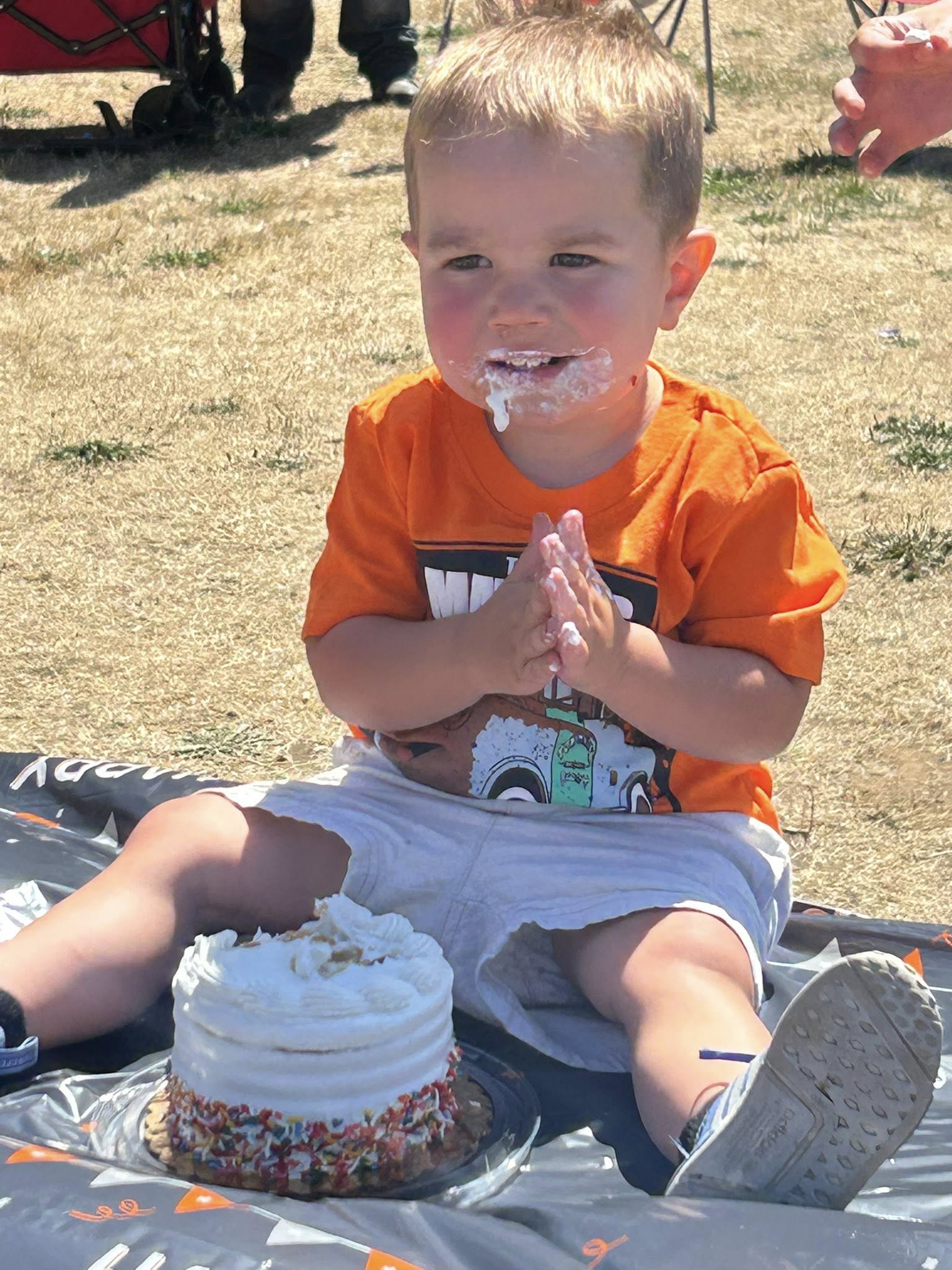 Toddler in an orange t-shirt clapping with frosting on his face next to a white birthday cake outdoors.