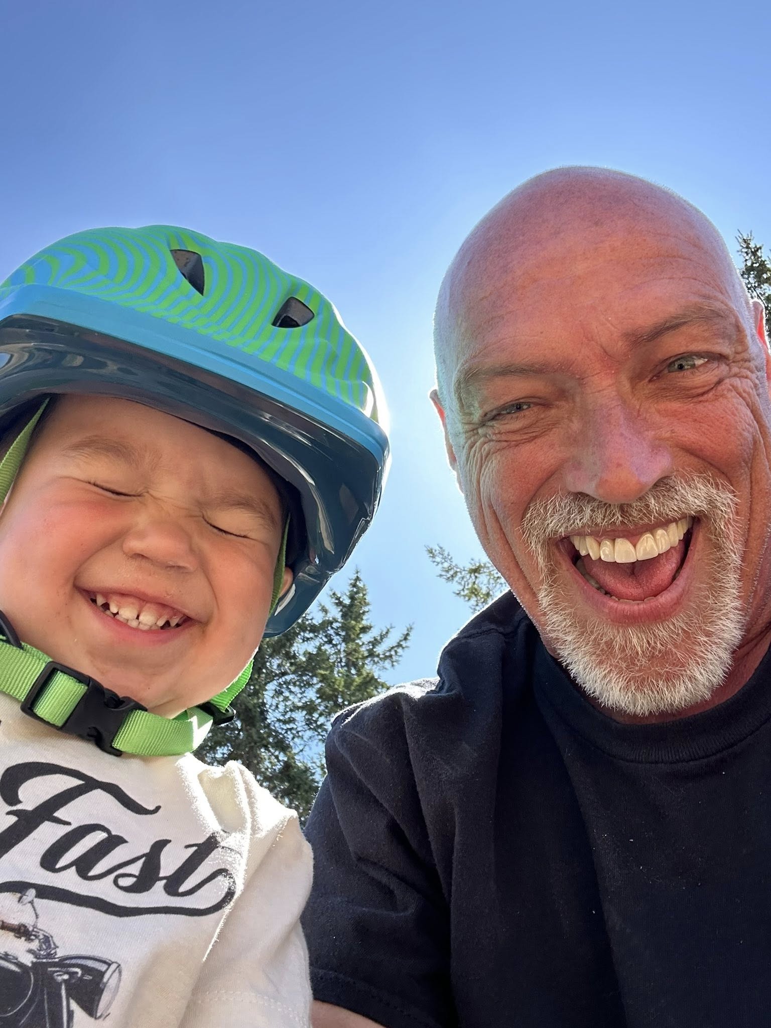 Toddler wearing a green helmet and an adult smiling together outdoors in the sun