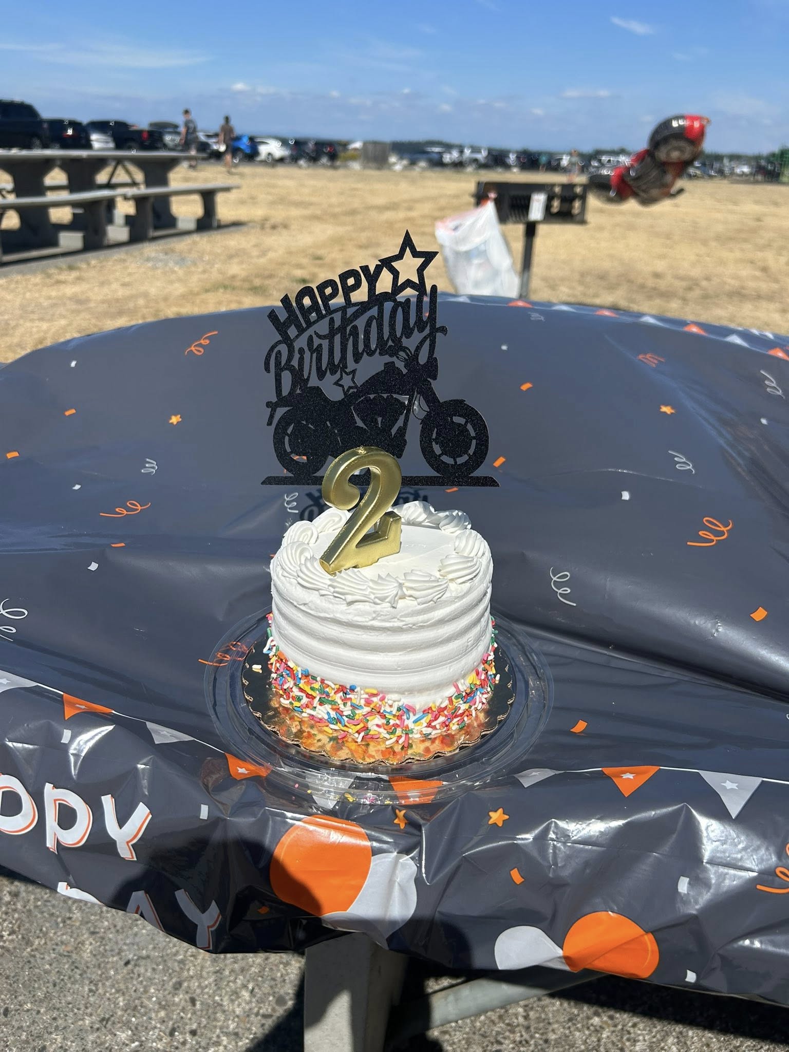 Black motorcycle-themed Happy Birthday cake topper with a gold number 2 candle on a white frosted birthday cake.