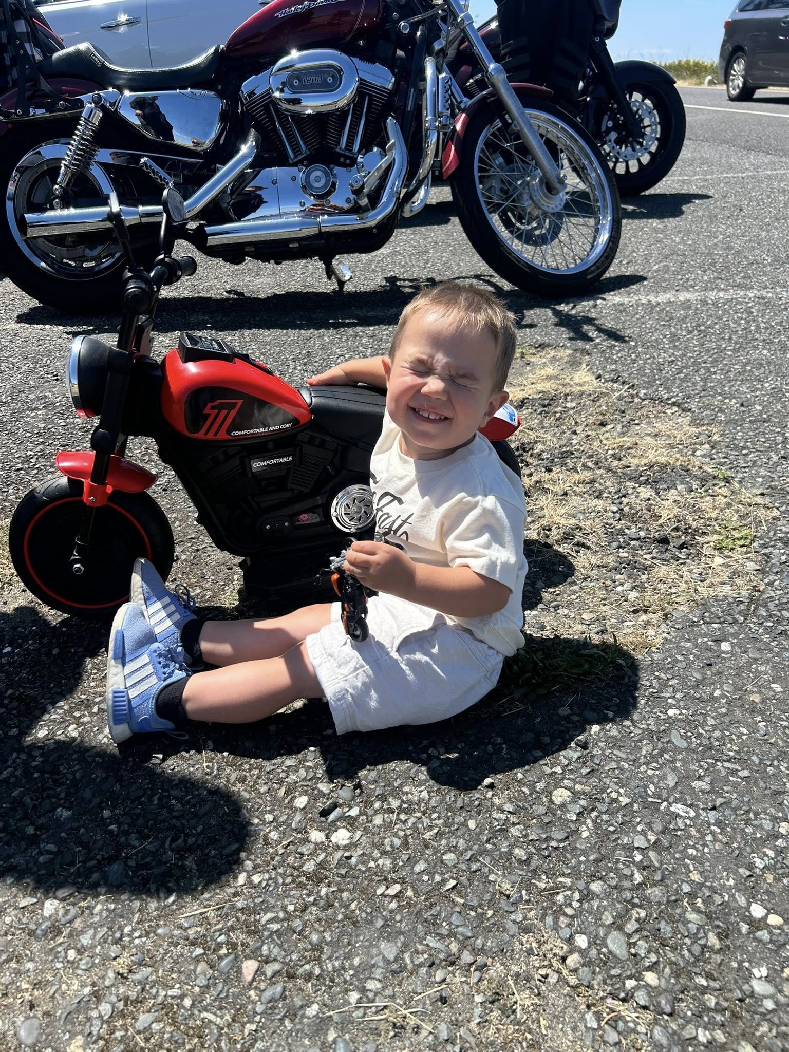 Toddler sitting on the ground next to a red toy motorcycle in a parking lot with a full-size motorcycle nearby.
