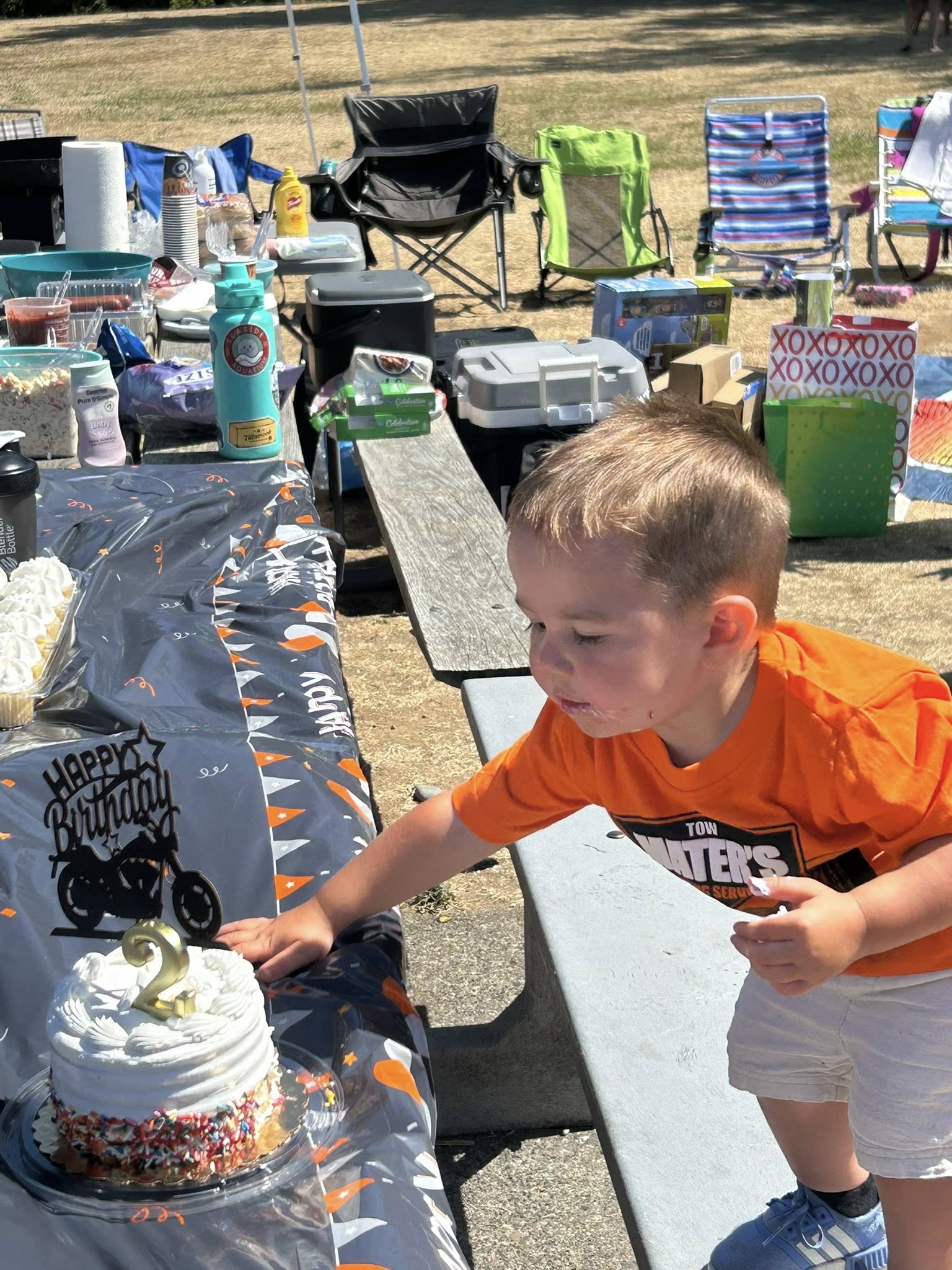 A young boy in an orange shirt reaching for his 2nd birthday cake decorated with a motorcycle topper.
