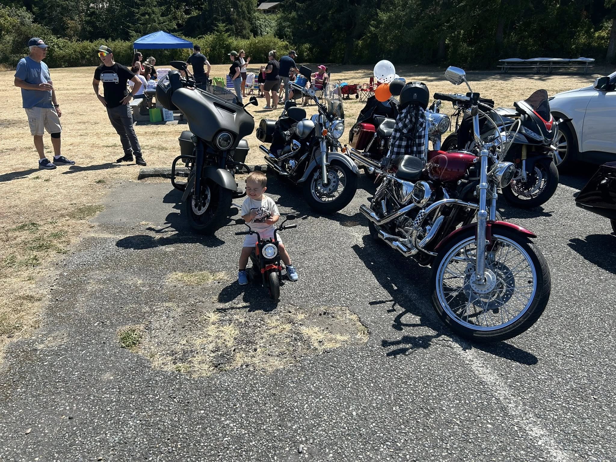 Toddler sitting on a small toy motorcycle parked alongside adult motorcycles at an outdoor event.