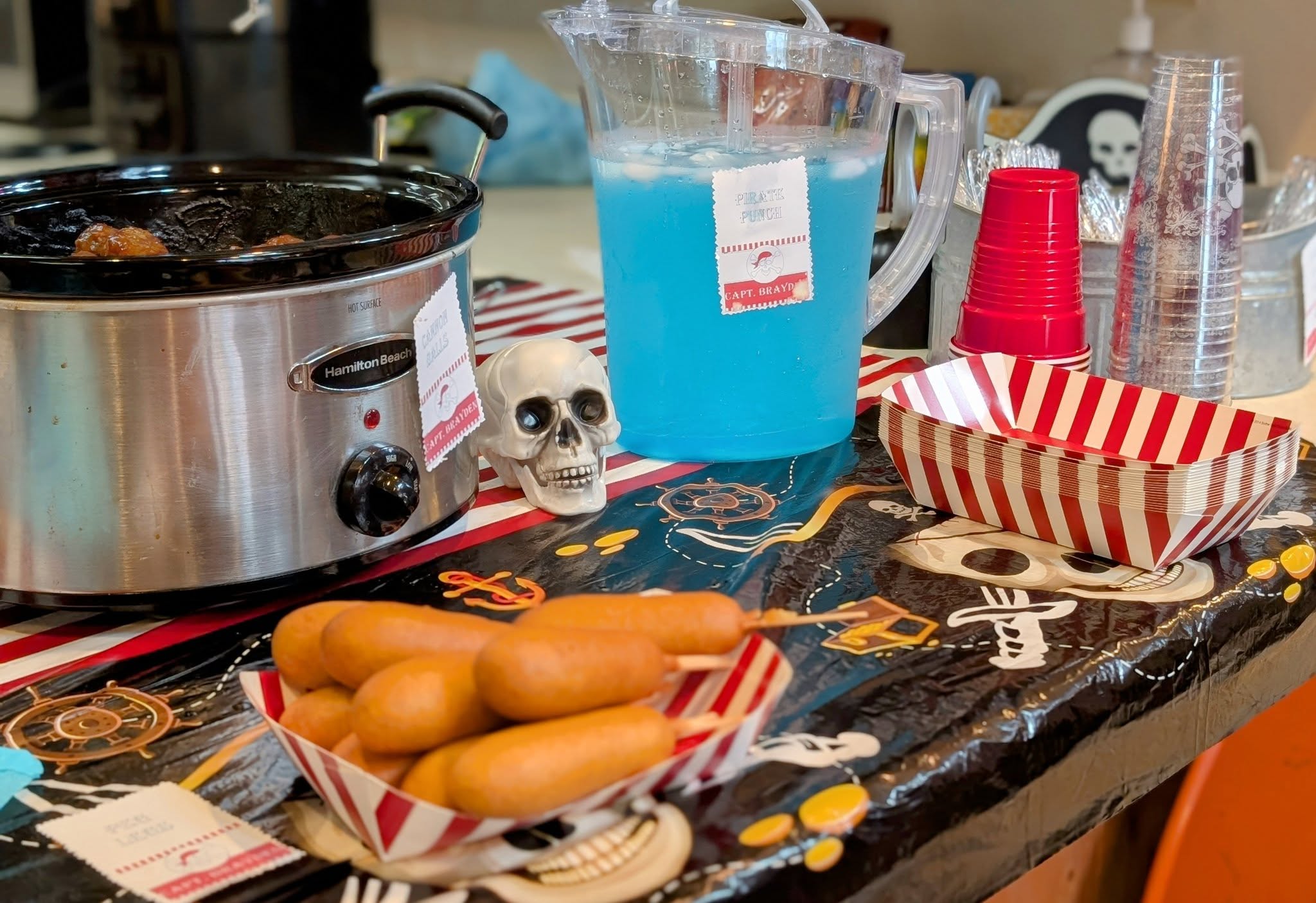 Pirate-themed party table featuring a crockpot, blue pirate punch, and corn dogs on a nautical tablecloth.
