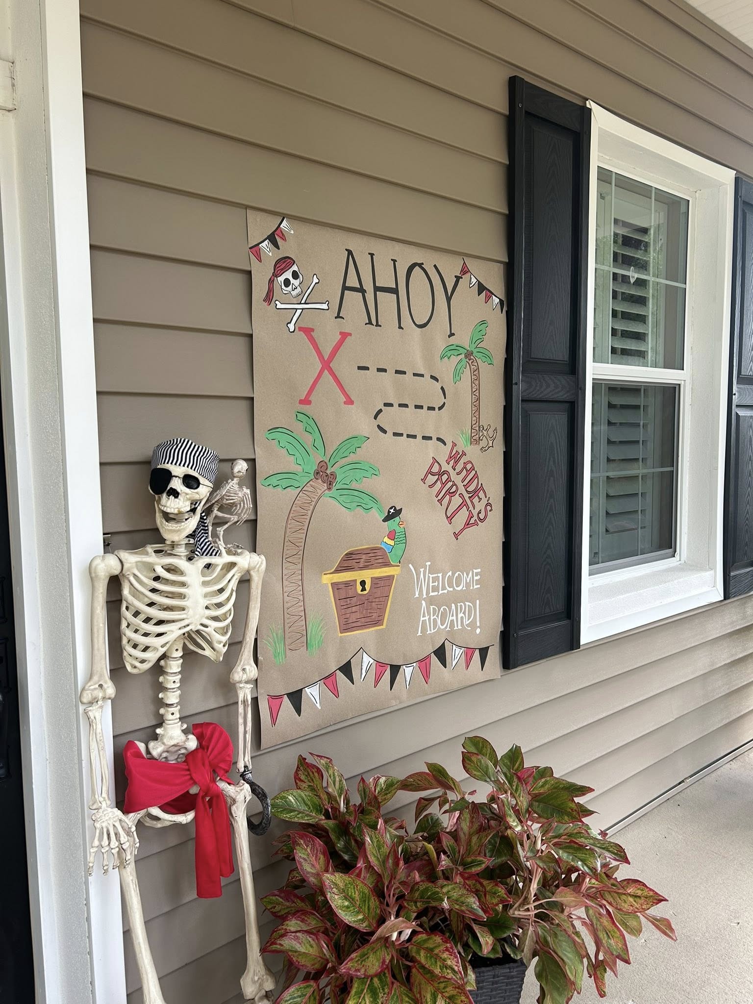 Skeleton wearing a pirate bandana greeting guests with an Ahoy pirate-themed party sign on a porch.