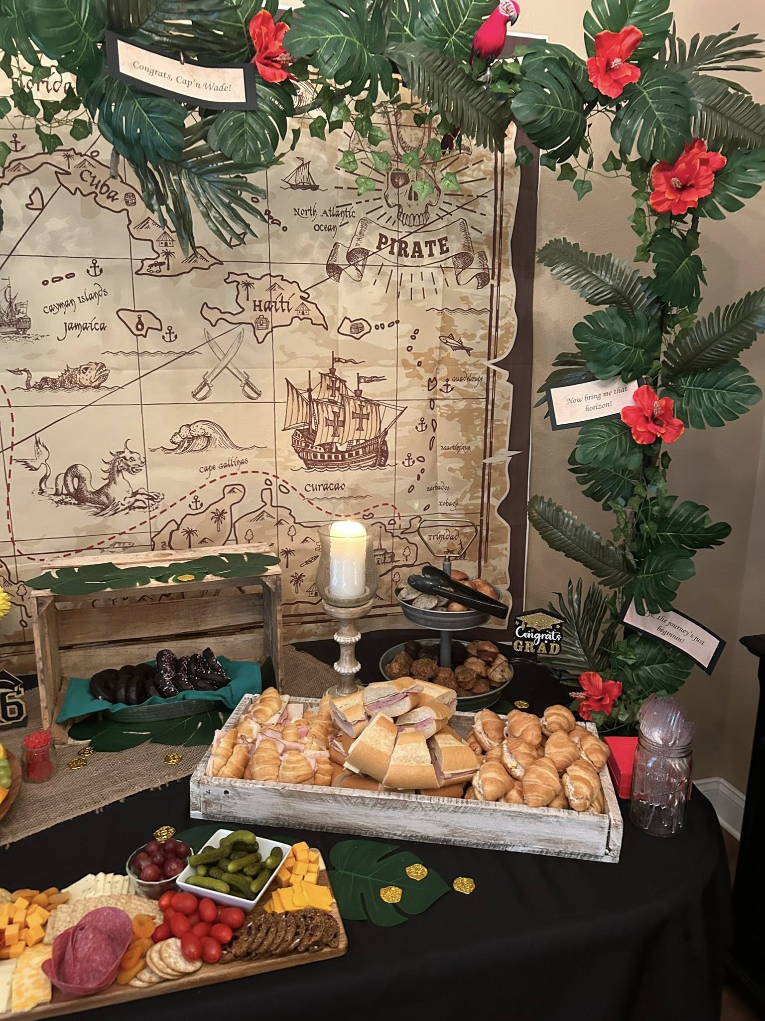 Pirate-themed graduation party food table with a treasure map backdrop, deli sandwiches, cheese, and cookies.