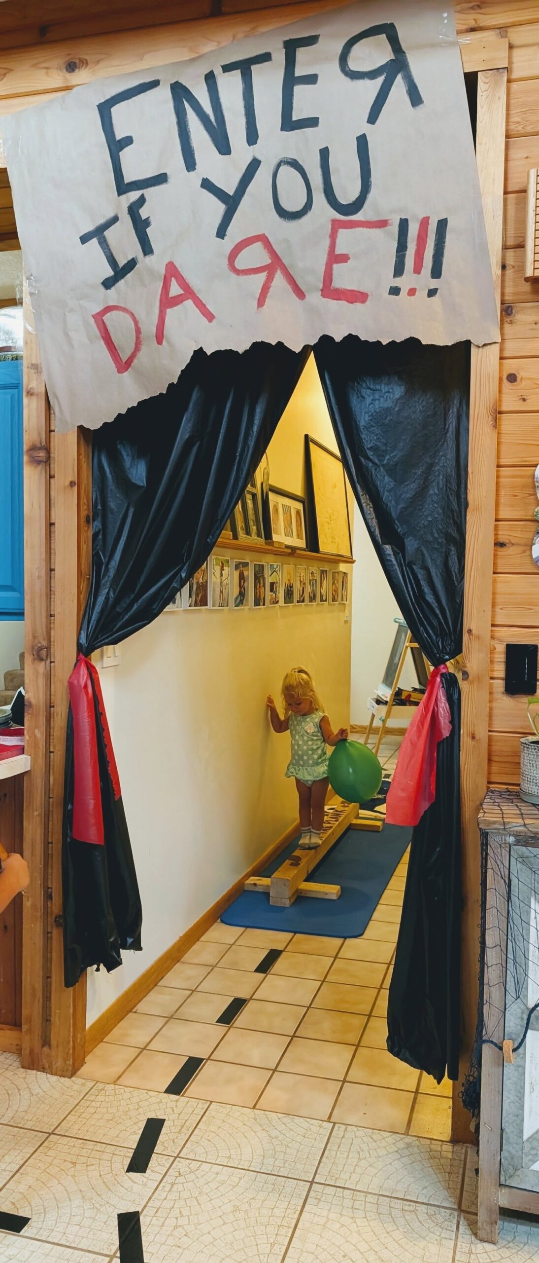 Toddler balancing on a wooden beam, holding a green balloon, in a doorway decorated with a sign that says enter if you dare.