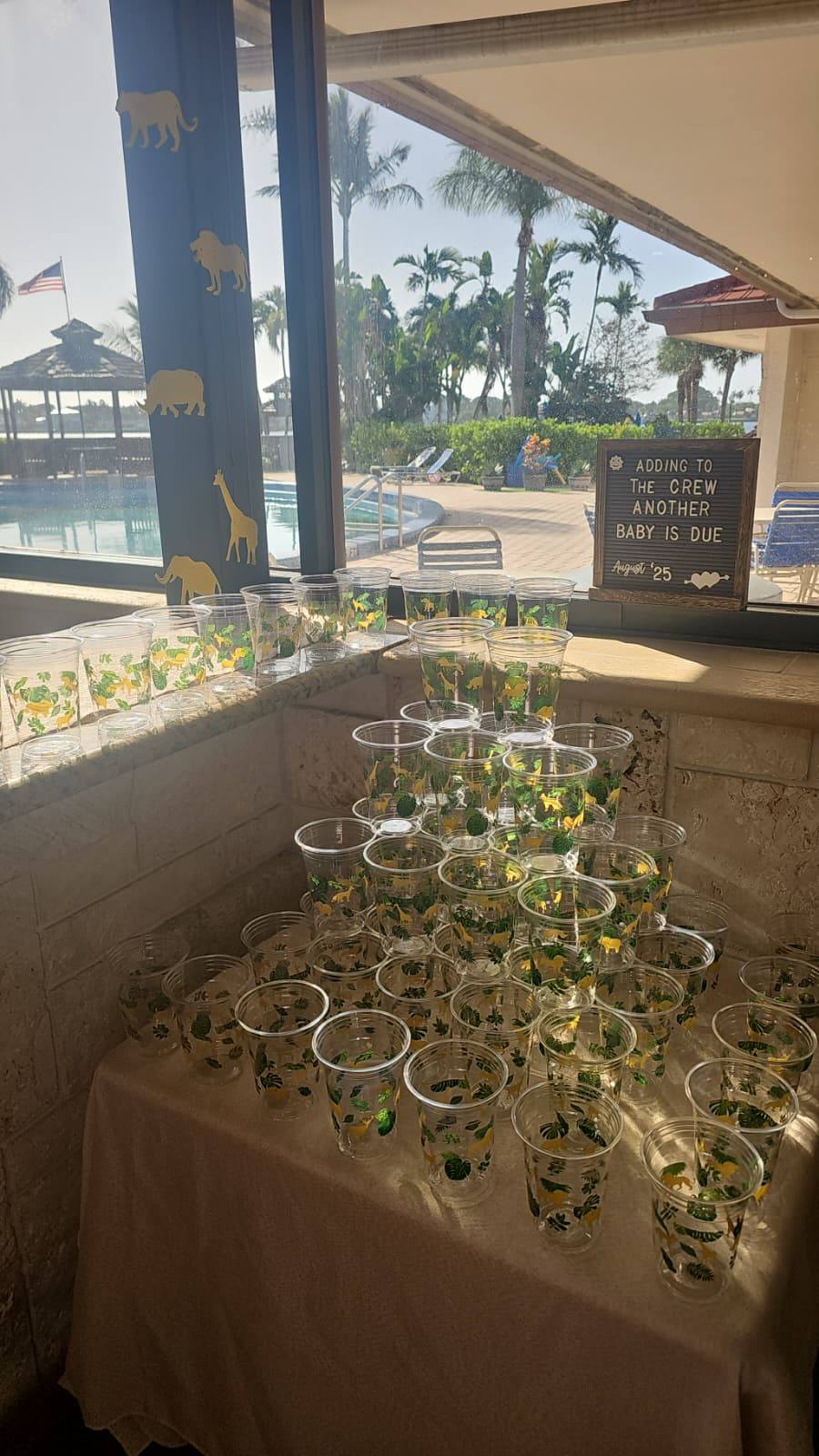 Stack of clear plastic cups with jungle animal print at a baby shower with a sign stating a baby is due August 25.