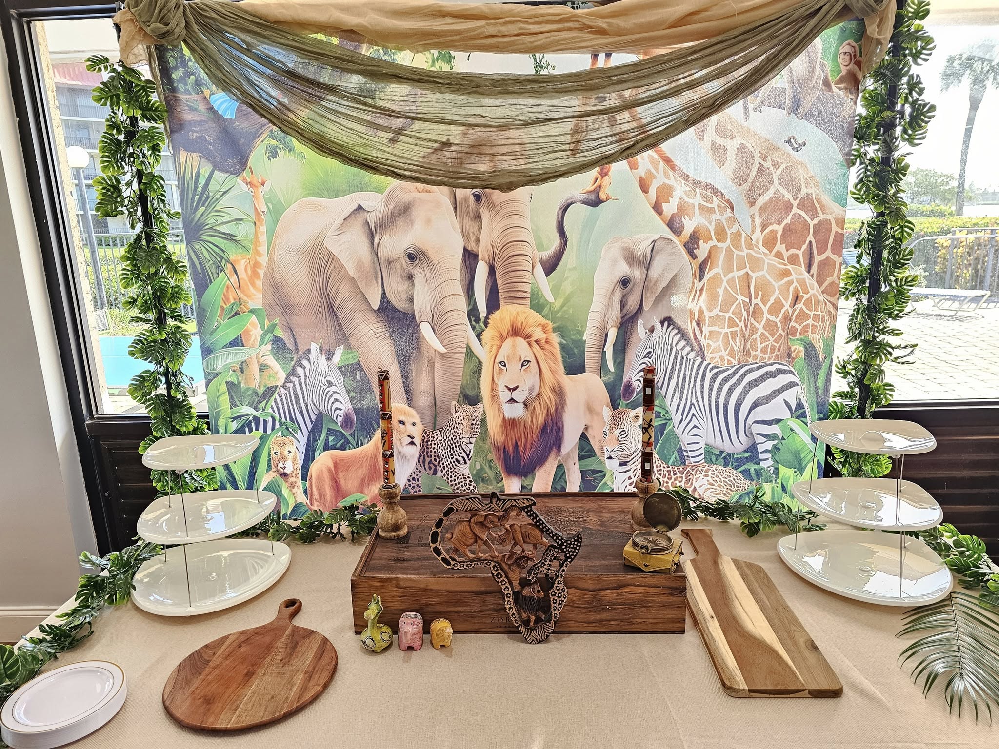Safari themed party table display with a backdrop of African wildlife, tiered snack stands, and wooden serving boards.