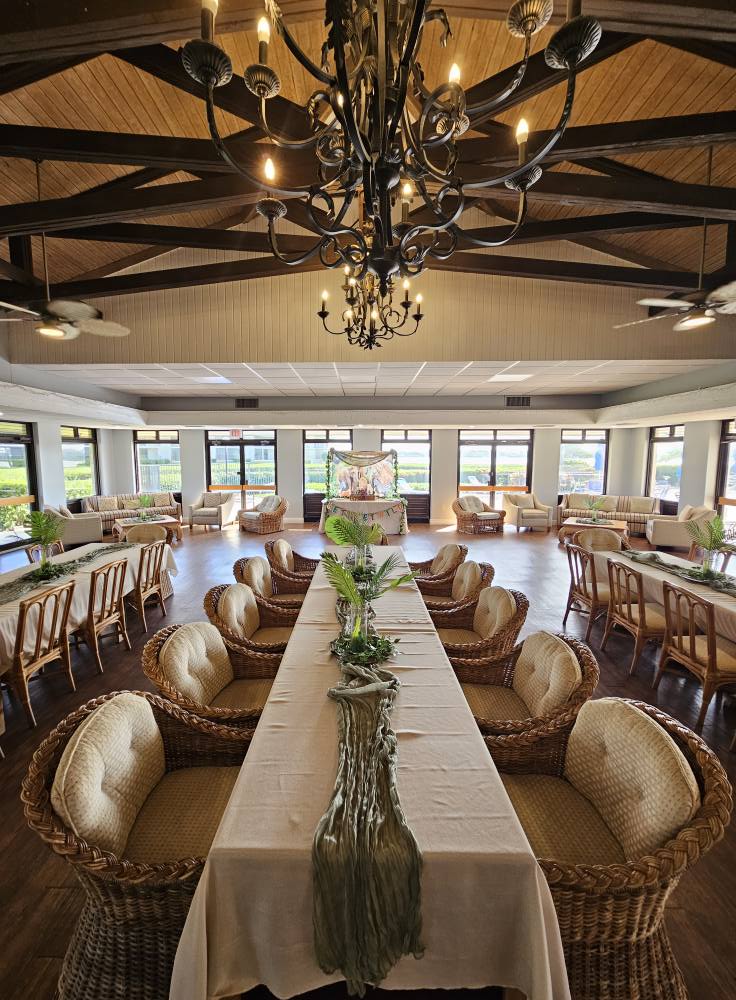 Dining room with wicker chairs and long tables arranged for an event beneath a large chandelier.