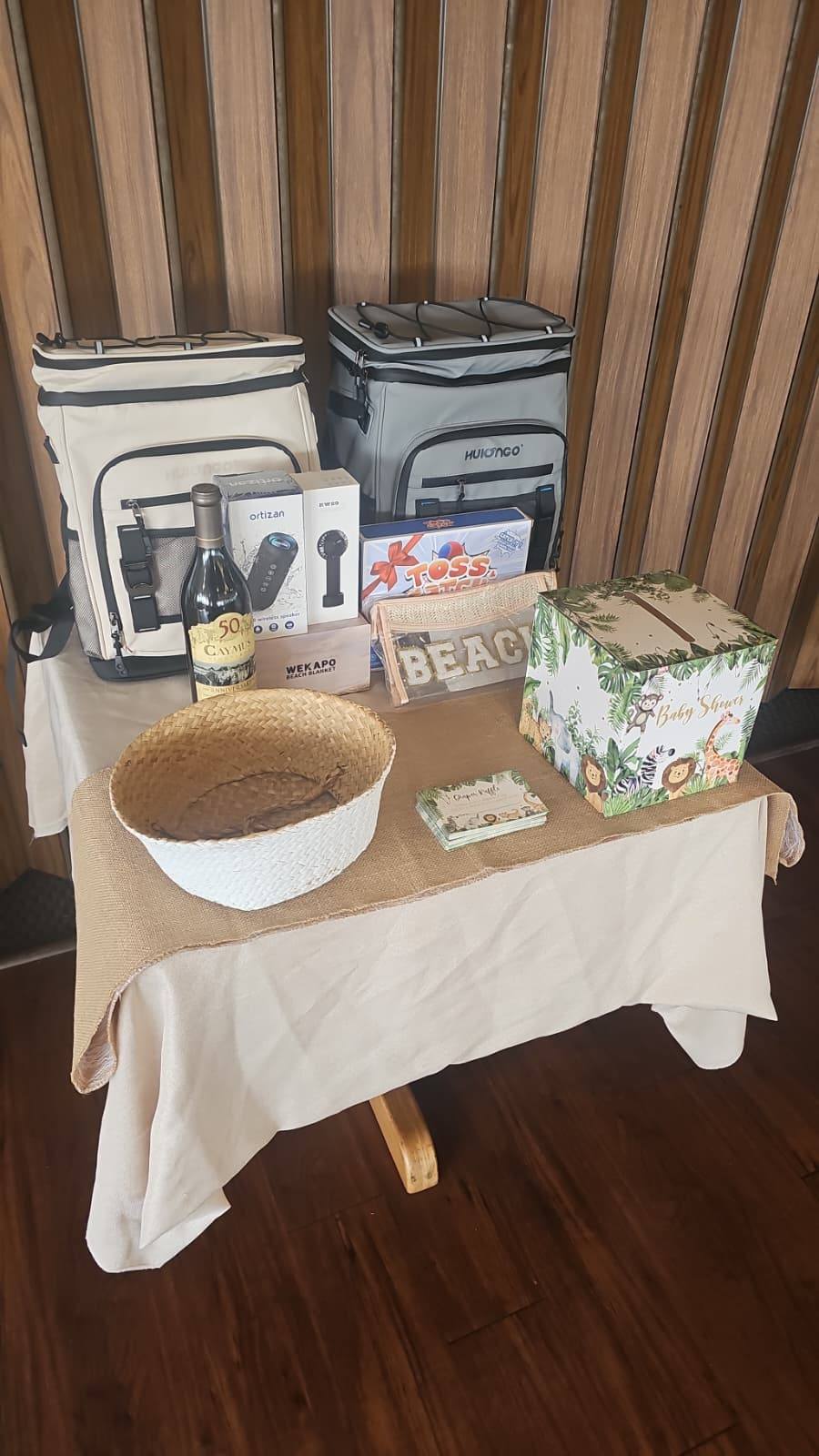 Display table featuring raffle prizes including Huiongo cooler backpacks, Caymus wine, and a baby shower card box.