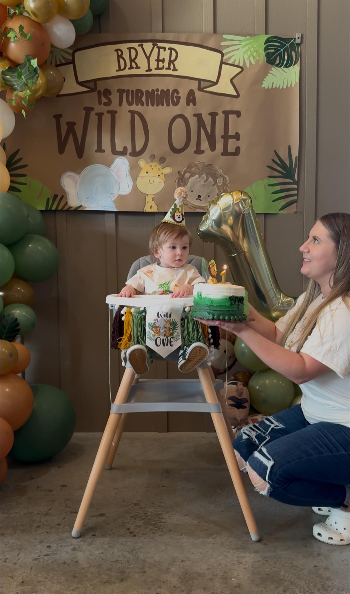 Toddler in a high chair wearing a party hat in front of a Wild One themed birthday backdrop with a smash cake.