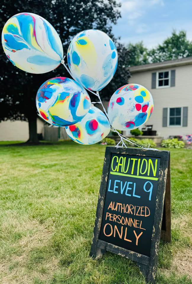 A-frame chalkboard sign reading Caution Level 9 Authorized Personnel Only with colorful marbled balloons tied to it in a yard.
