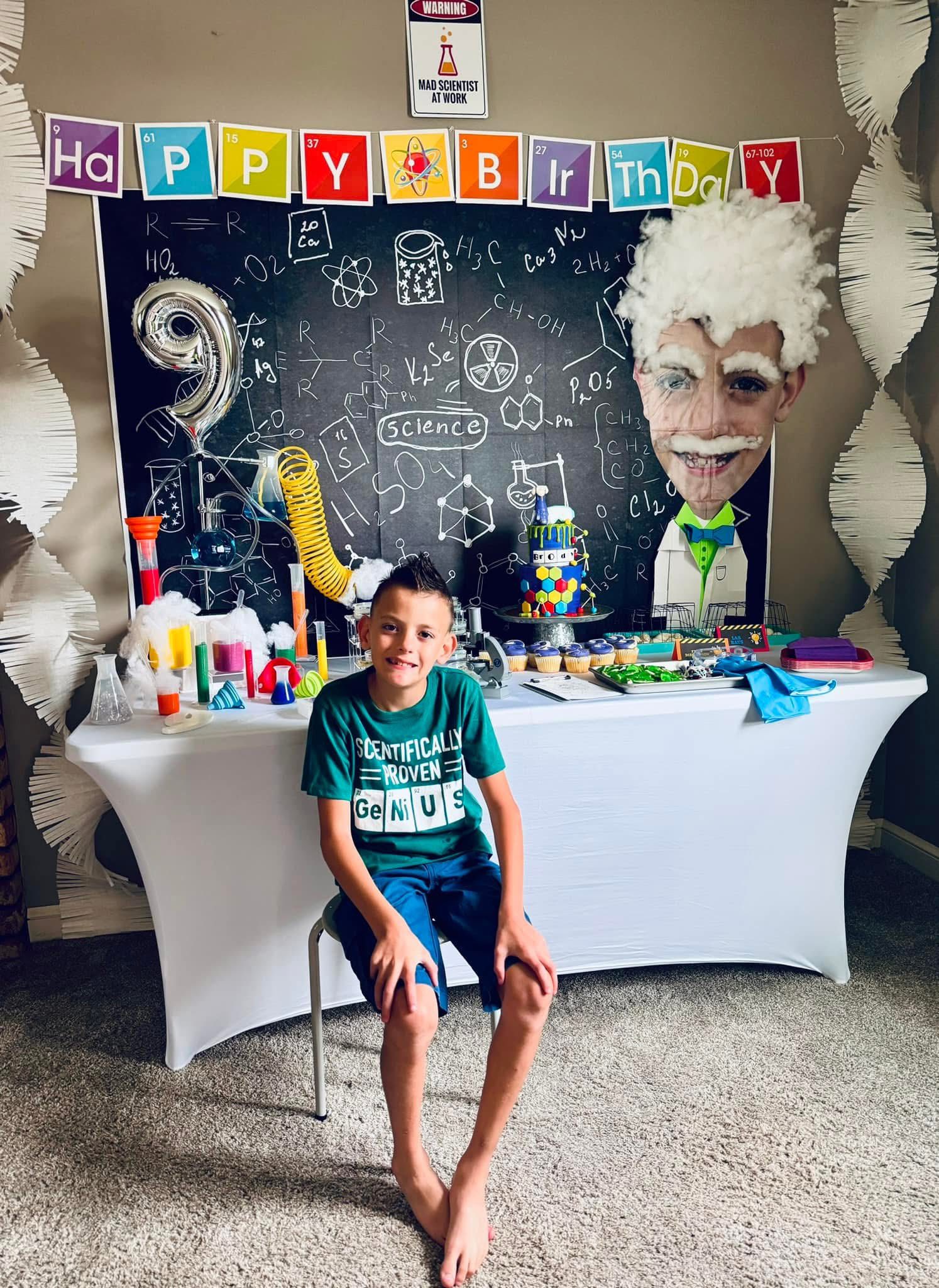 A young boy sits in front of a table decorated for a science-themed 9th birthday party.