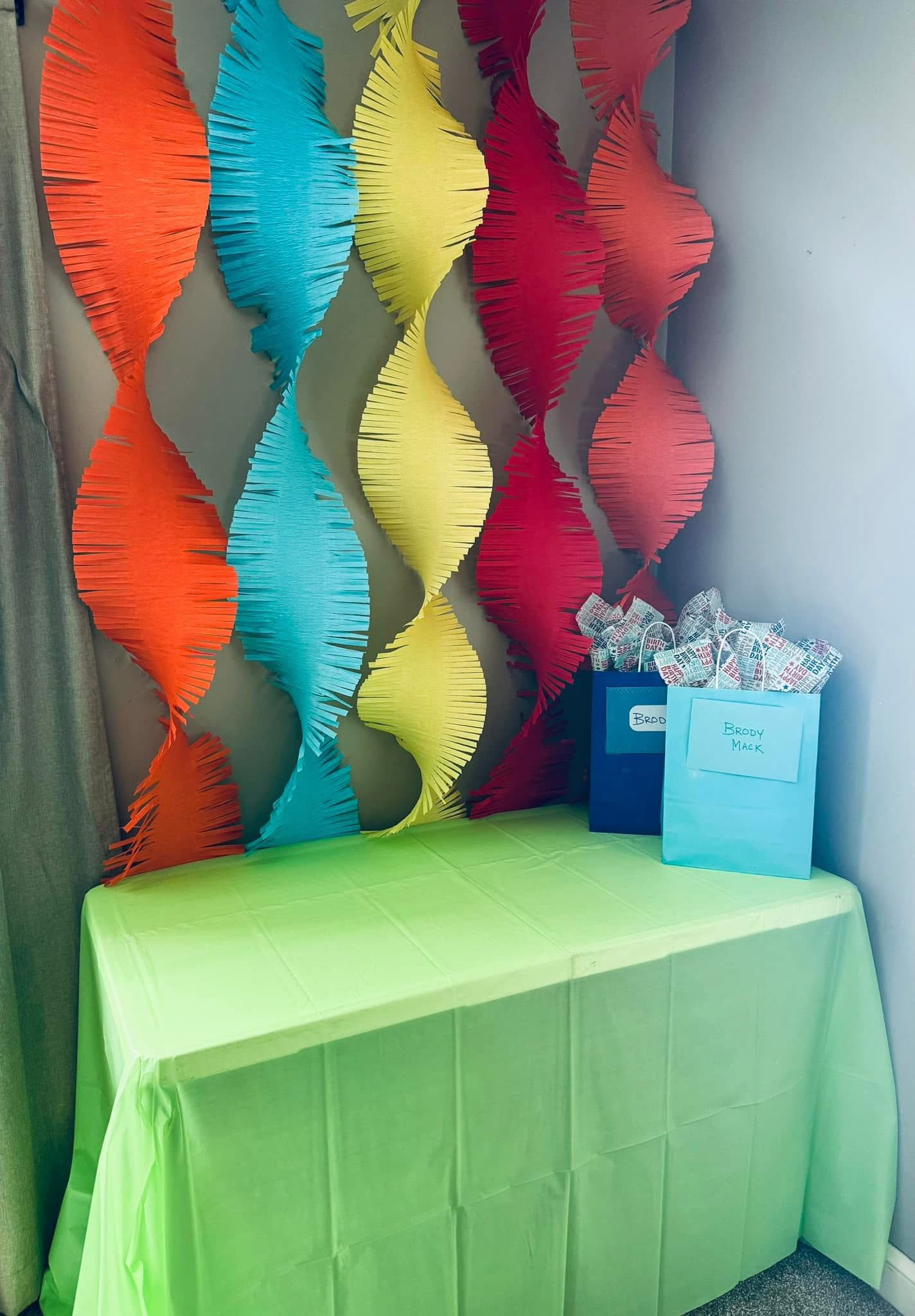 Table with lime green tablecloth, gift bags, and colorful hanging paper decorations behind it.