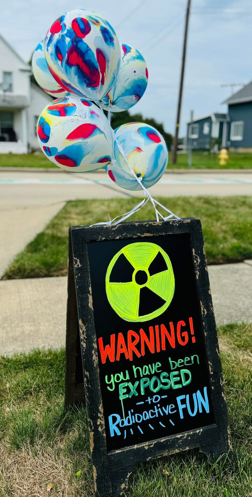 A sidewalk chalkboard sign topped with colorful tie-dye balloons reading Warning: You have been exposed to Radioactive FUN.