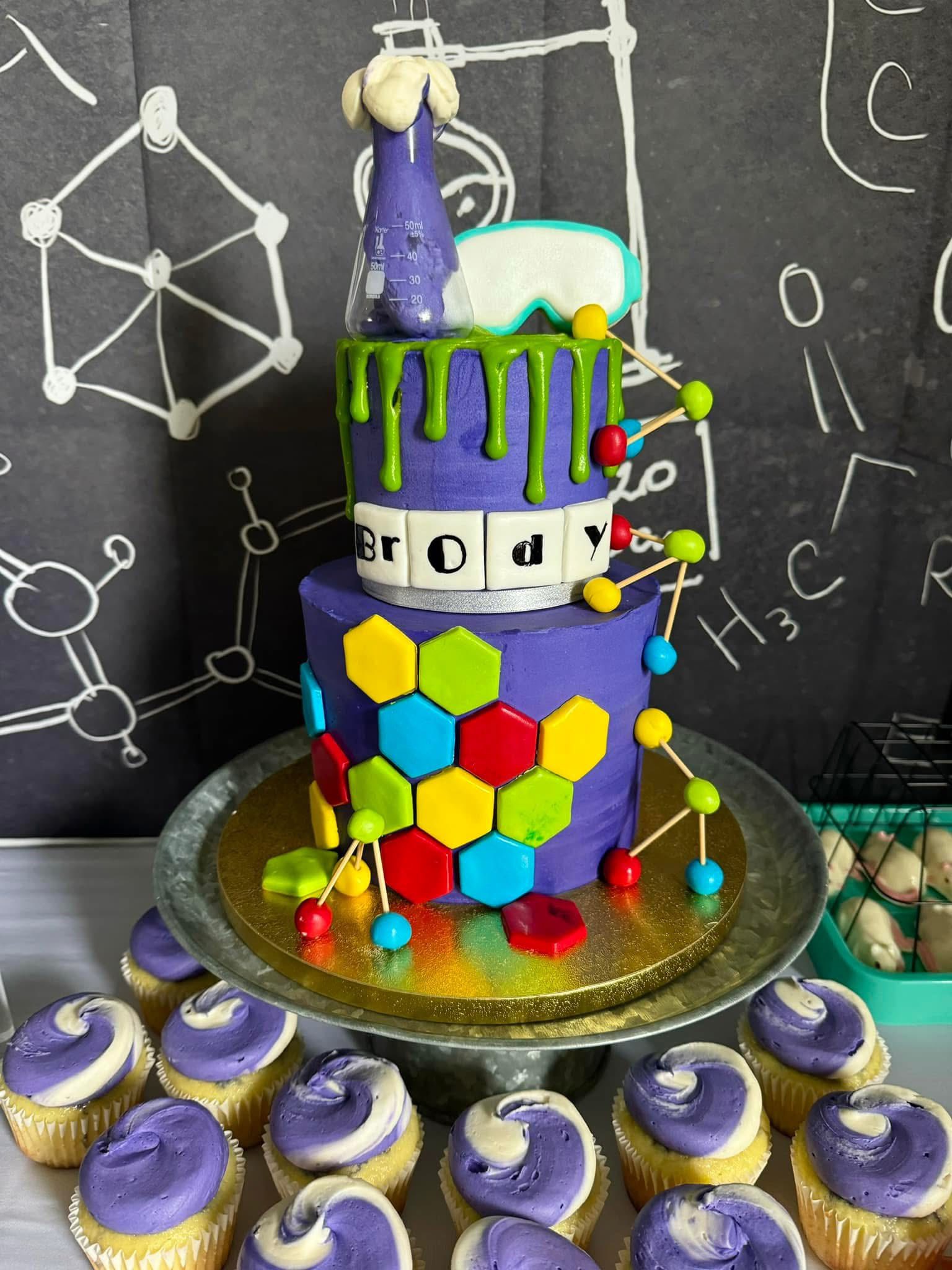 Two-tier purple science-themed birthday cake decorated with colorful hexagons, chemistry beakers, and name blocks.