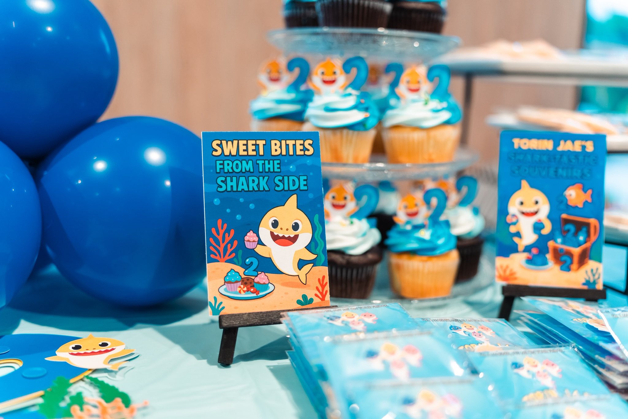 Shark-themed table sign reading Sweet Bites From The Shark Side with cupcakes in the background.