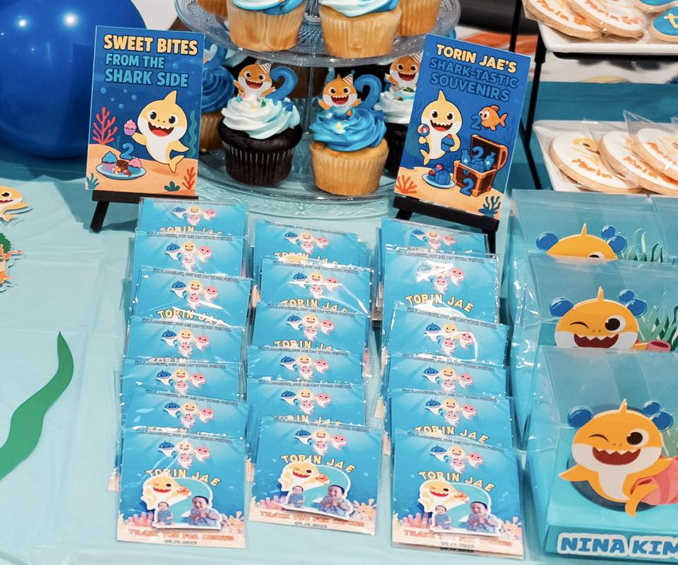 Table display featuring Baby Shark themed party favors, cupcakes, and personalized thank you cards for a 2nd birthday.