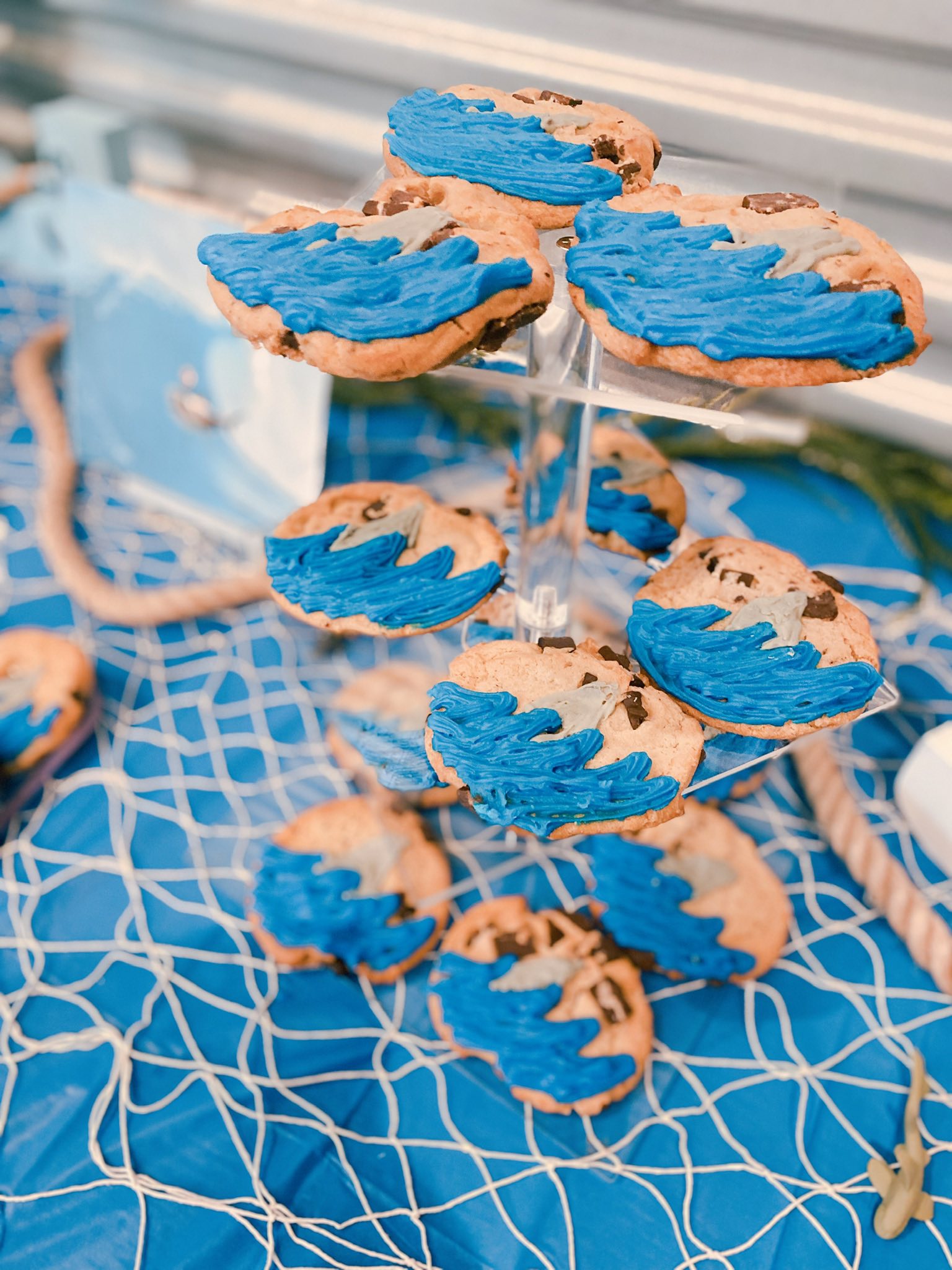 Chocolate chip cookies decorated with blue wave frosting, stacked on a tiered stand at a nautical-themed party.