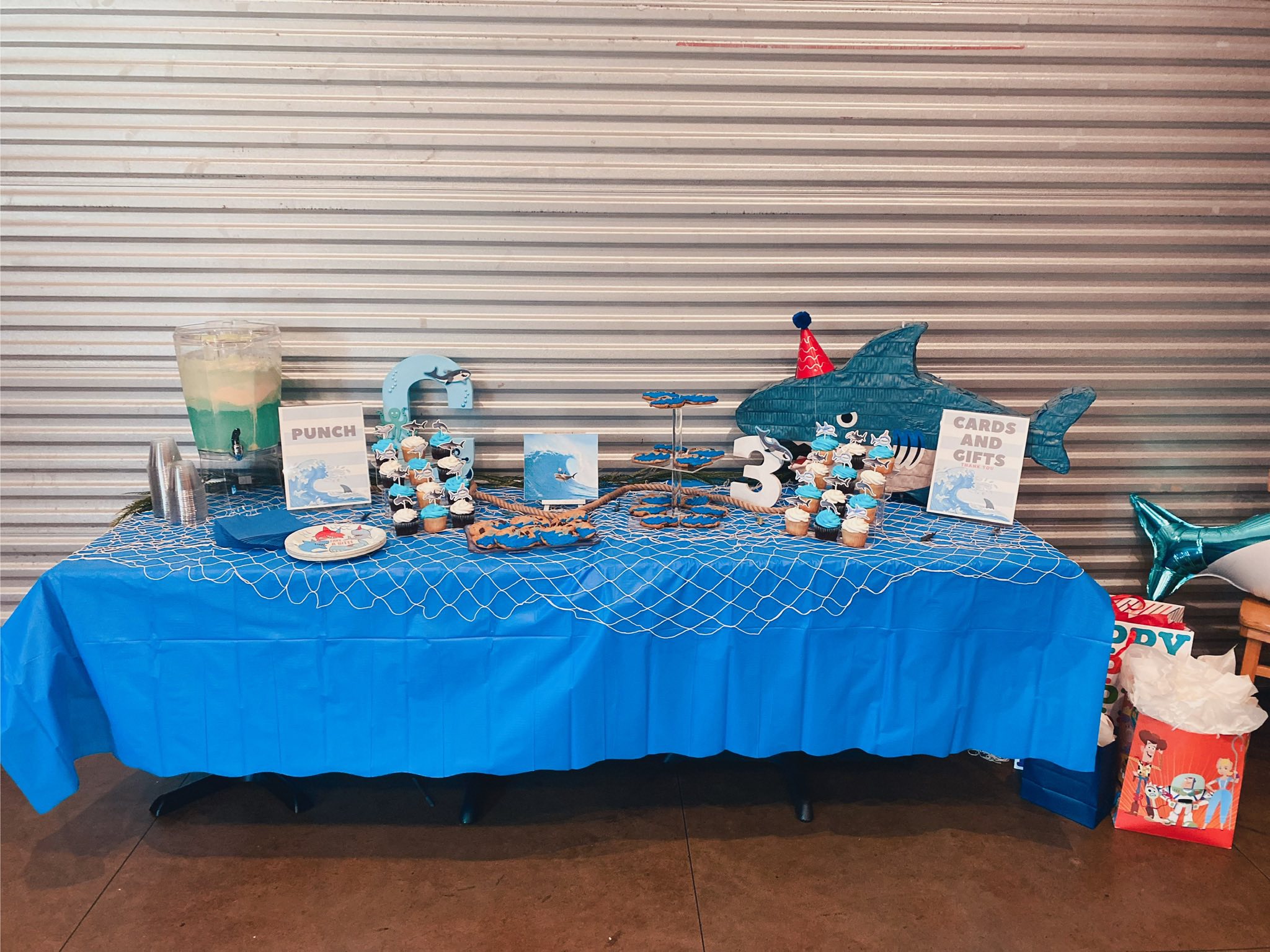 Shark-themed birthday party table with cupcakes, punch, card station, and shark pinata