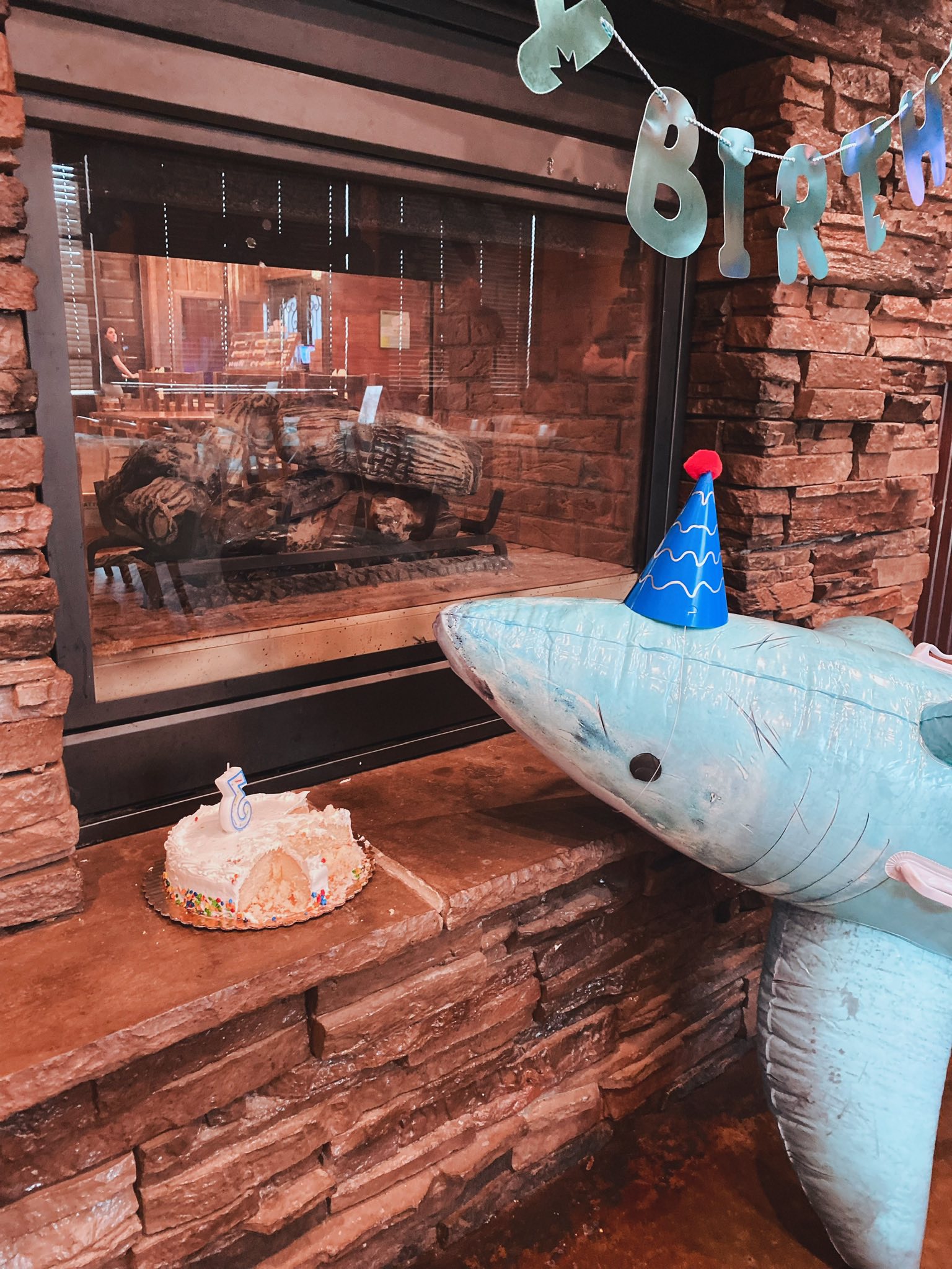 Inflatable shark wearing a blue party hat next to a small birthday cake with a number 5 candle on a stone hearth.
