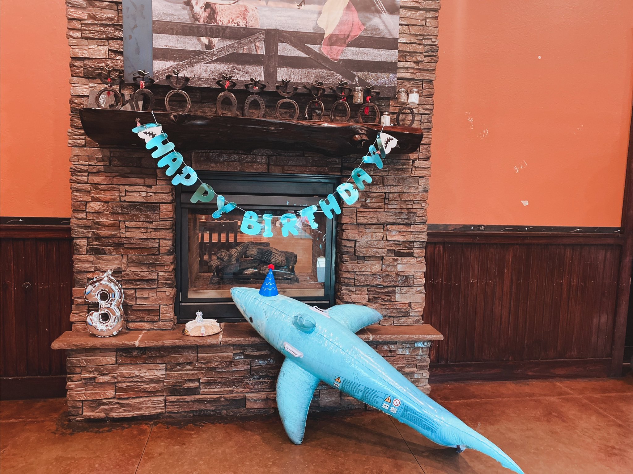 Inflatable shark wearing a party hat next to a number three balloon and a small birthday cake on a stone fireplace.