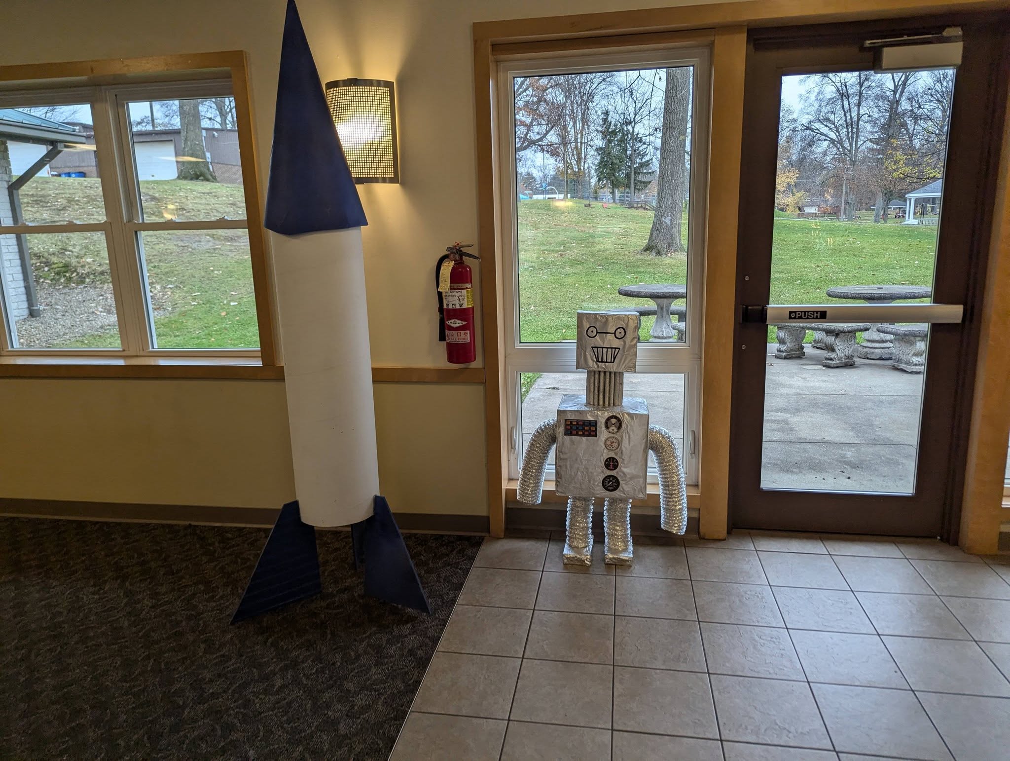 DIY paper rocket and silver robot props standing near a sliding glass door.