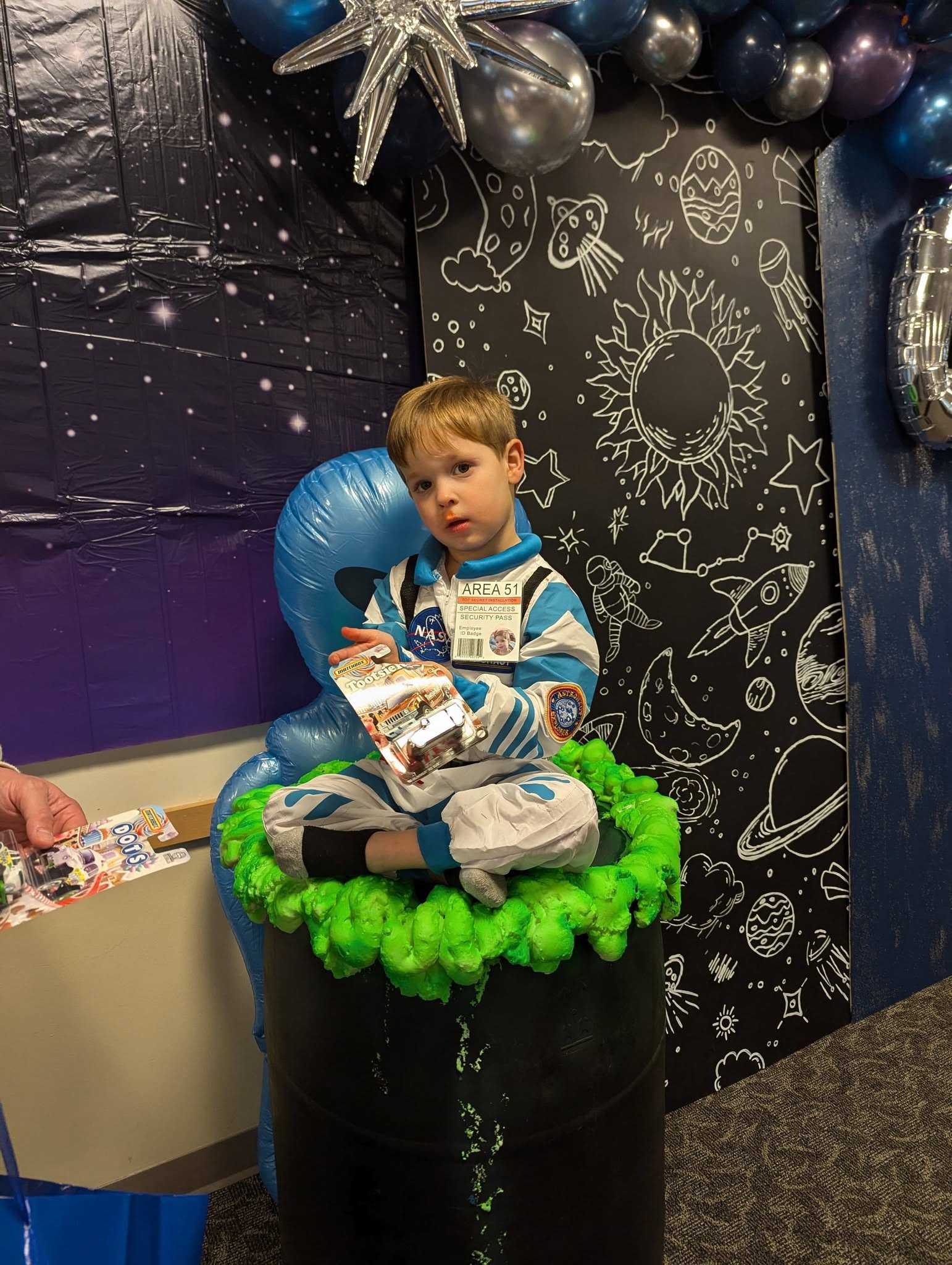 A young boy in an astronaut costume sitting in a barrel with green foam at an outer space themed birthday party.