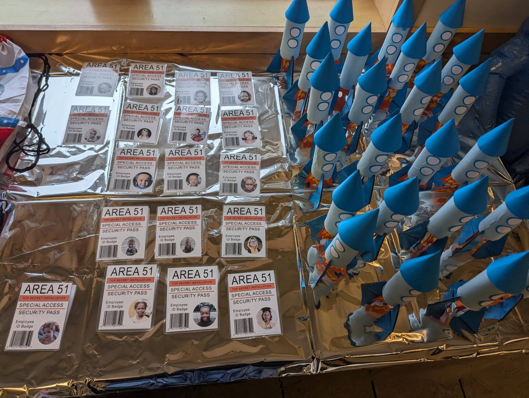 Handmade Area 51 security ID badges and paper toy rockets arranged on a silver surface for a party or event.