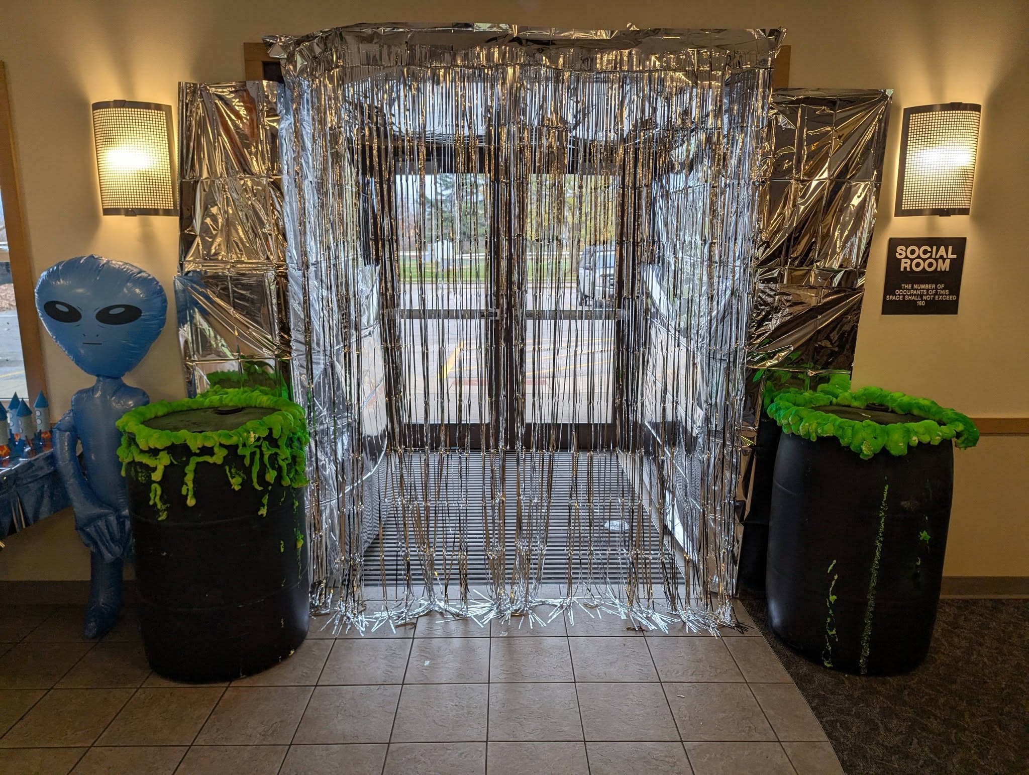 Entryway decorated for space theme with silver metallic fringe curtains, an inflatable alien, and toxic waste prop barrels.
