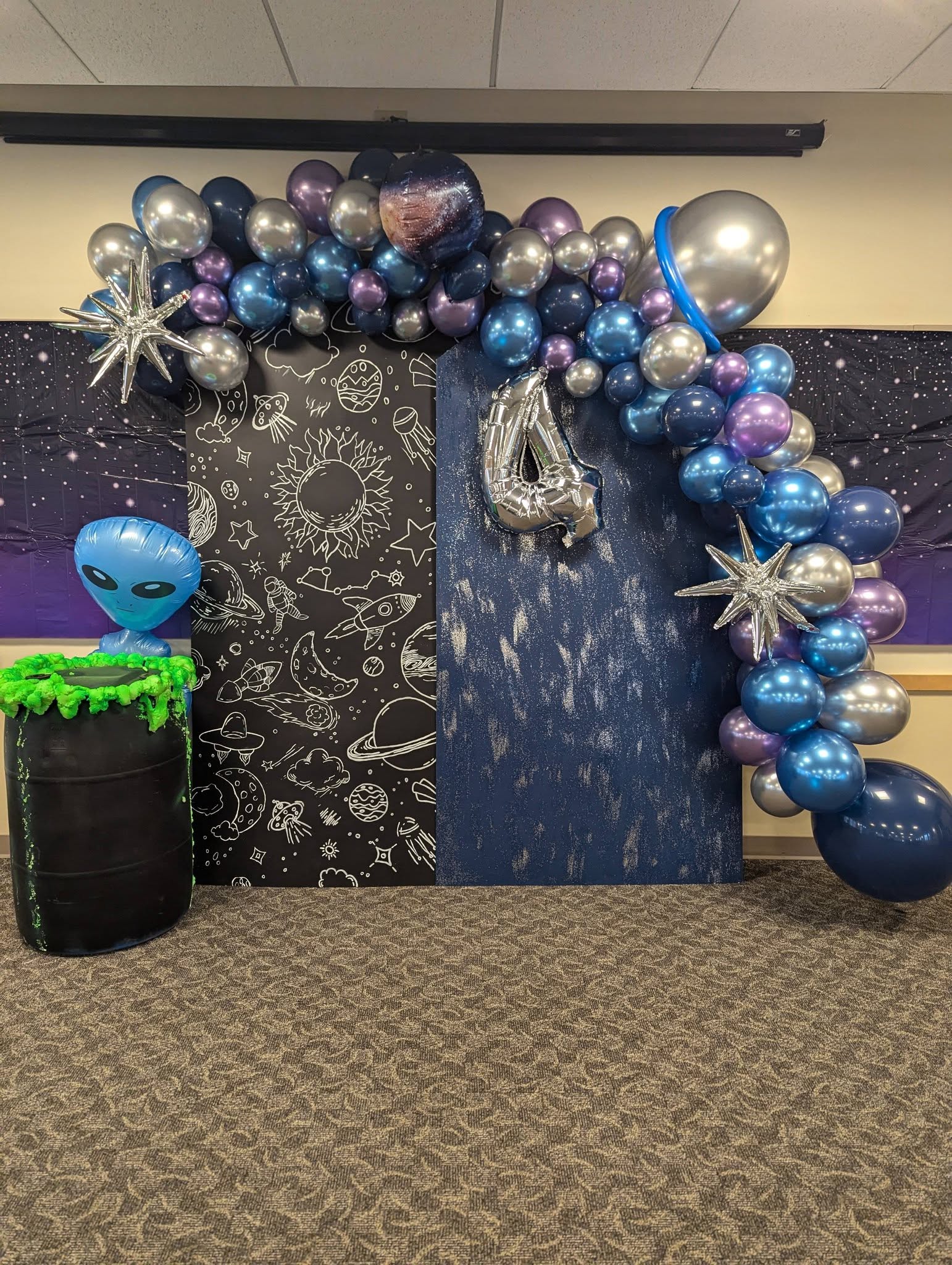 Space-themed party photo backdrop with rocket ship and alien balloons, glittery blue wall, and oversized number 4 foil balloon.