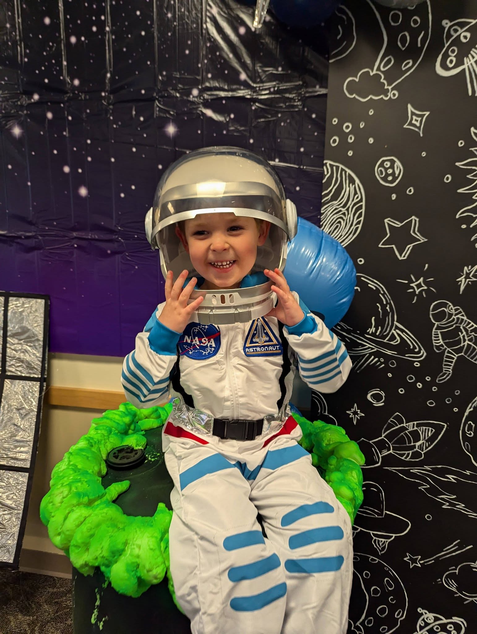 Young child wearing a white NASA astronaut costume with a helmet, sitting by space-themed decorations.