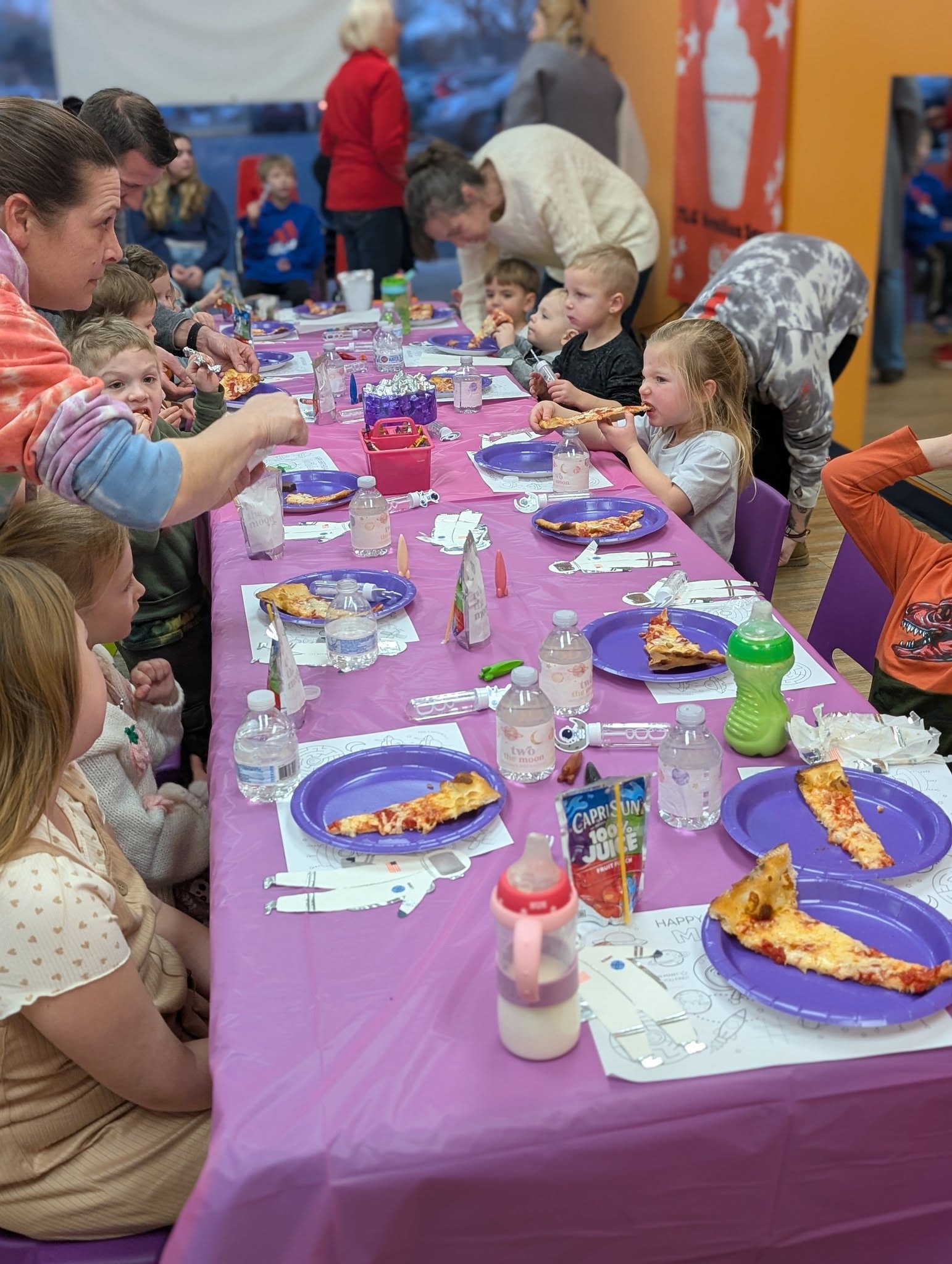 Children eating pizza at a long party table with purple tablecloths and space-themed decorations.