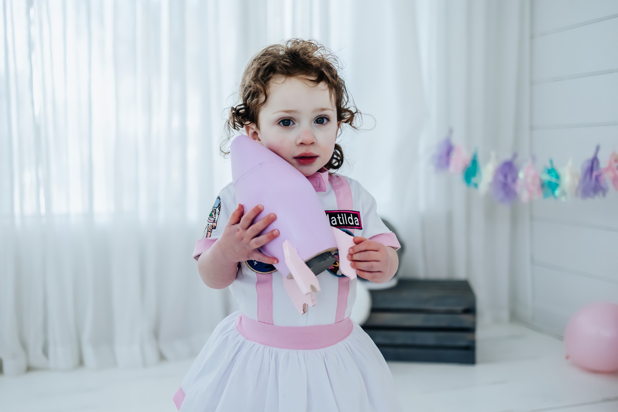Toddler wearing a custom white and pink astronaut dress with a personalized badge holding a model rocket ship.