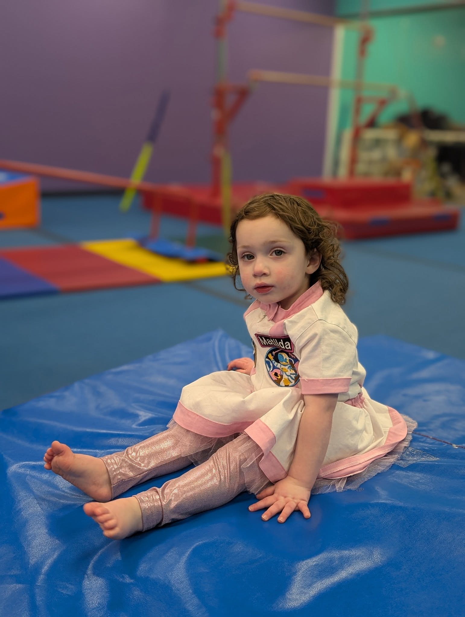Toddler in a white and pink dress with glittery leggings sitting on a blue gymnastics mat