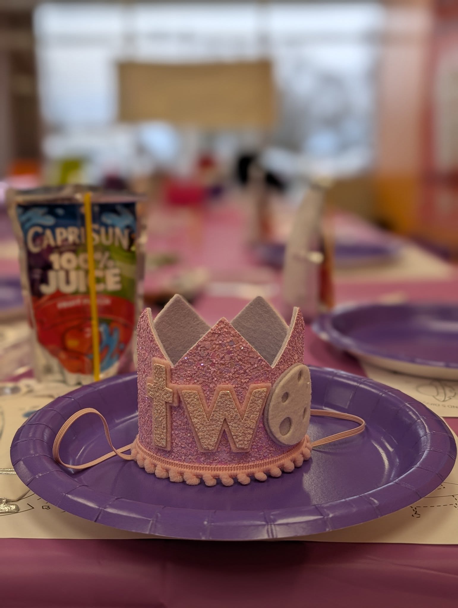 A pink glittery toddler birthday crown with the word 'two' on a purple party plate at a themed celebration.