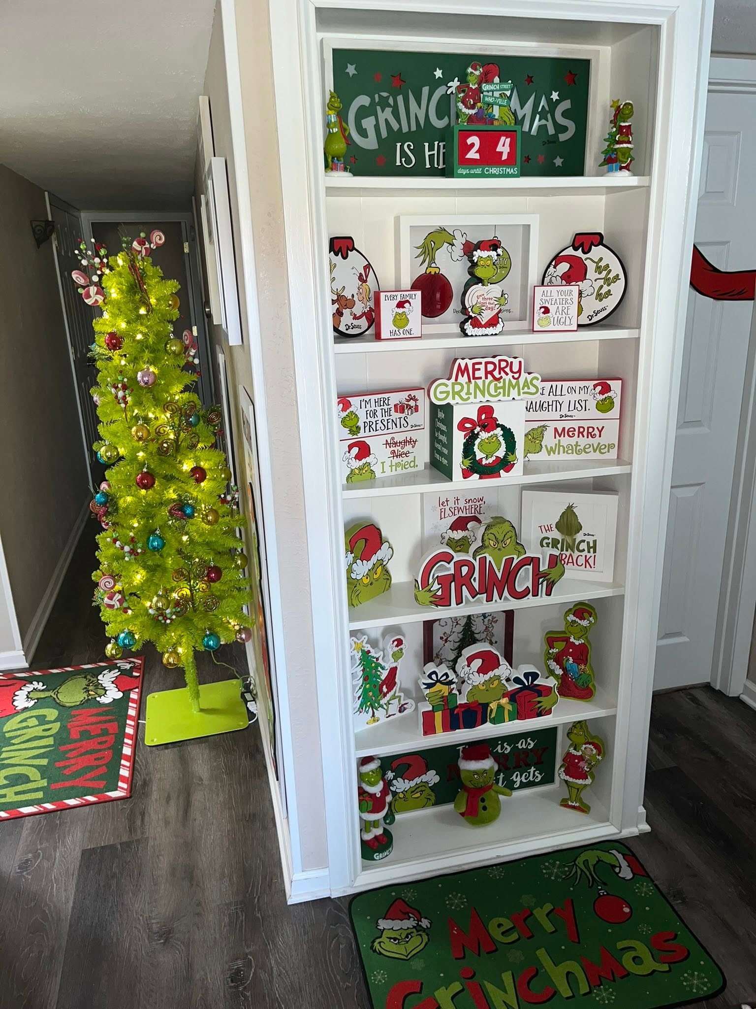 Shelves decorated with Grinch-themed Christmas signs, figures, and a bright green lighted Christmas tree nearby.