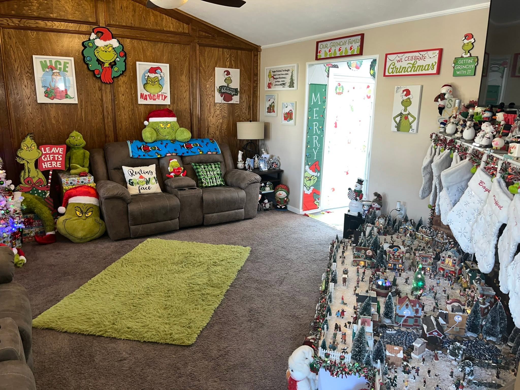 Living room filled with Grinch-themed Christmas decor, including plushies, signs, and a detailed holiday village.