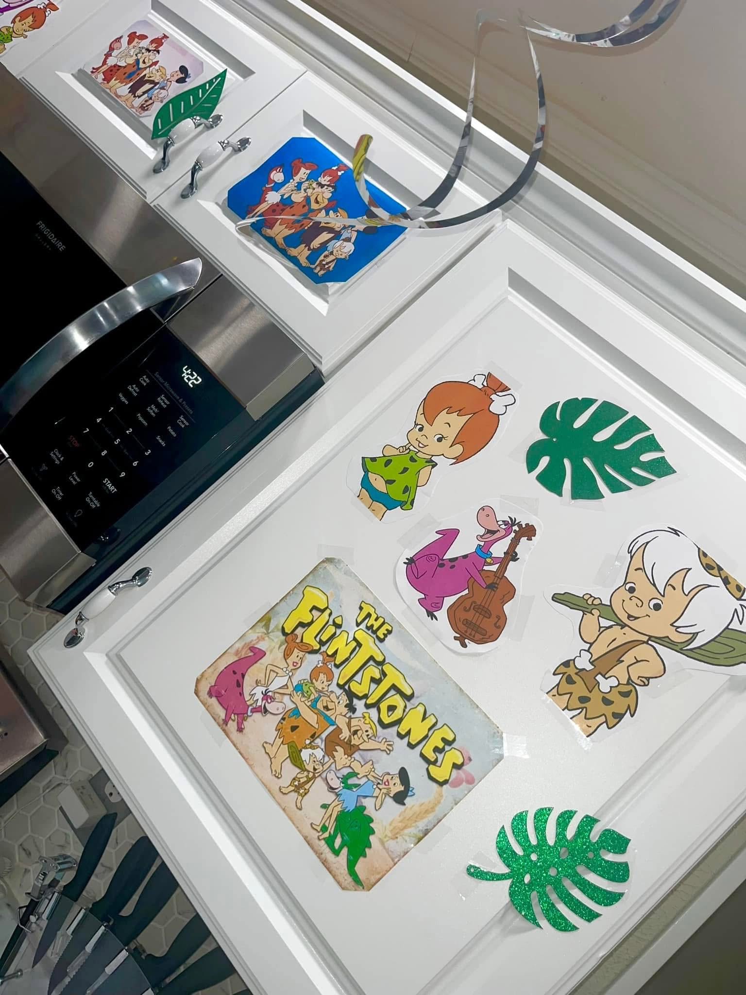 White kitchen cabinets decorated with Flintstones character decals and green monstera leaf cutouts.