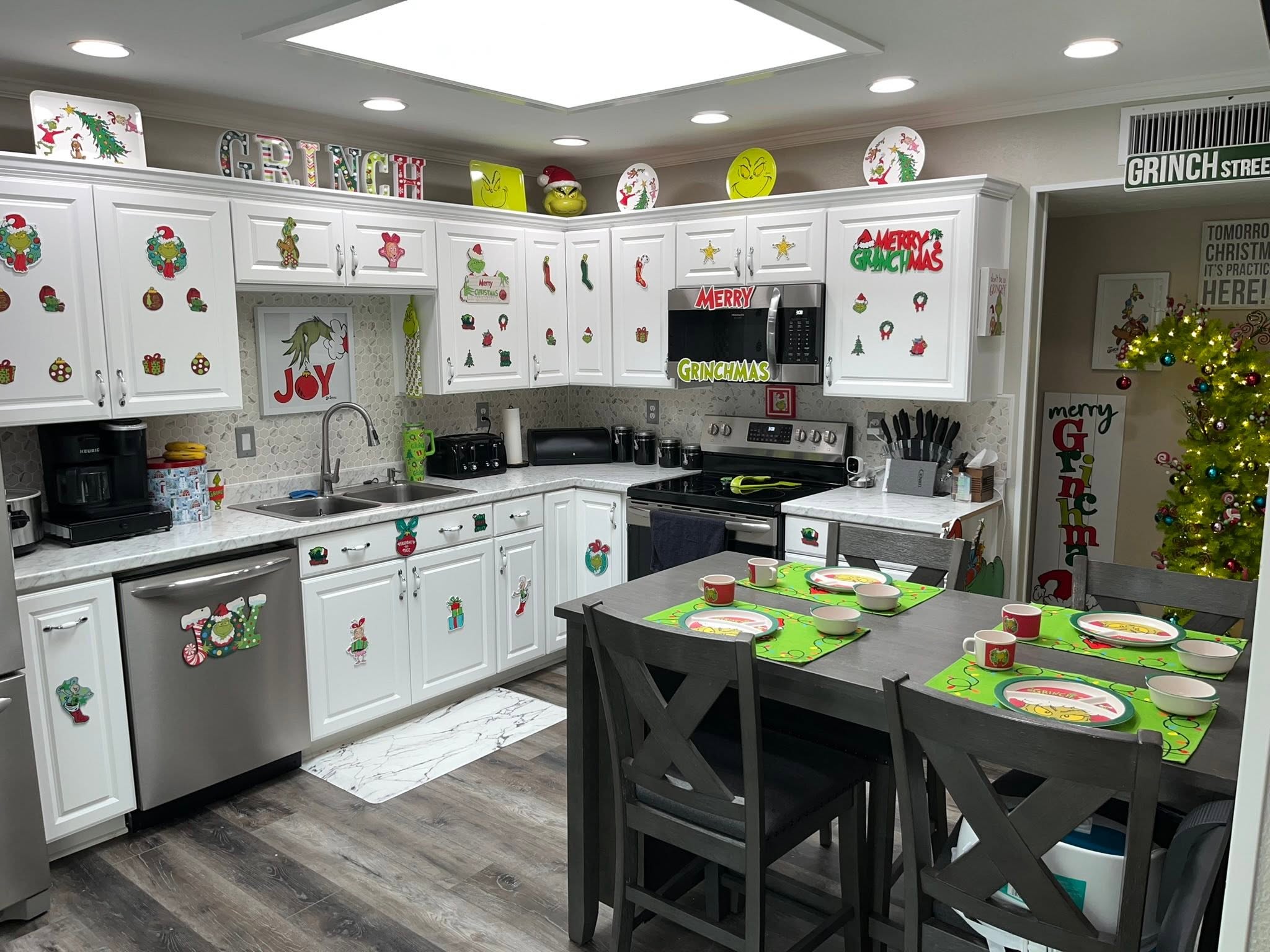 Kitchen decorated for Christmas with Grinch-themed decals on white cabinets, festive plates, and a decorated tree.