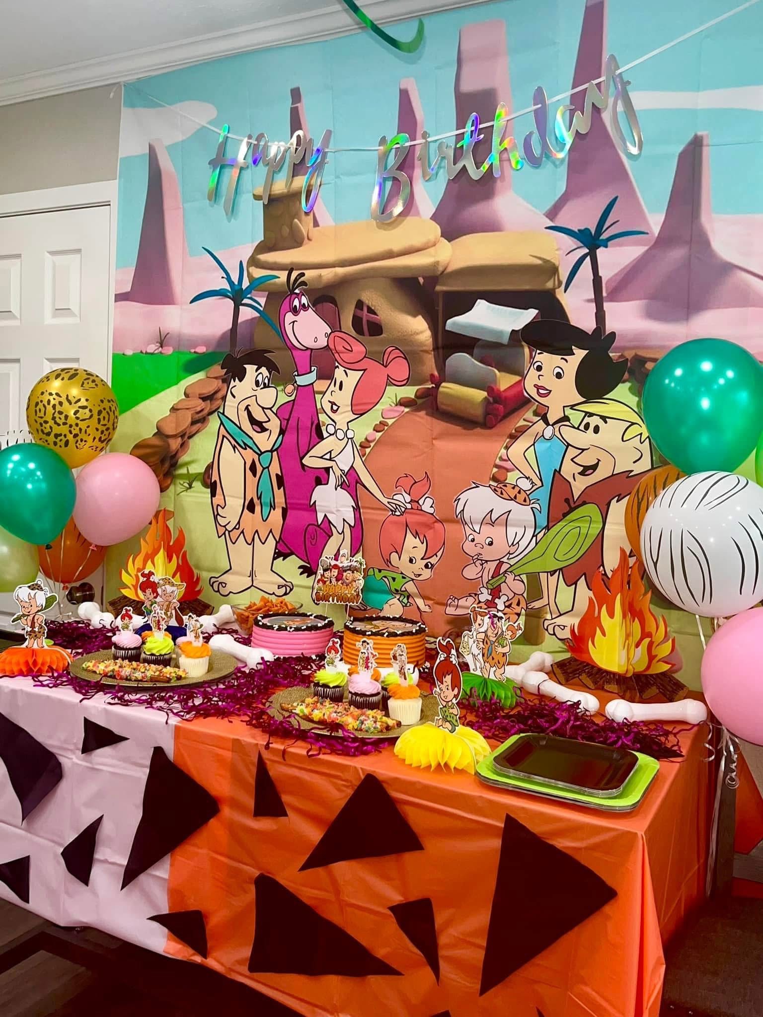 Flintstones themed birthday party table with cartoon character decorations, balloons, and treats.