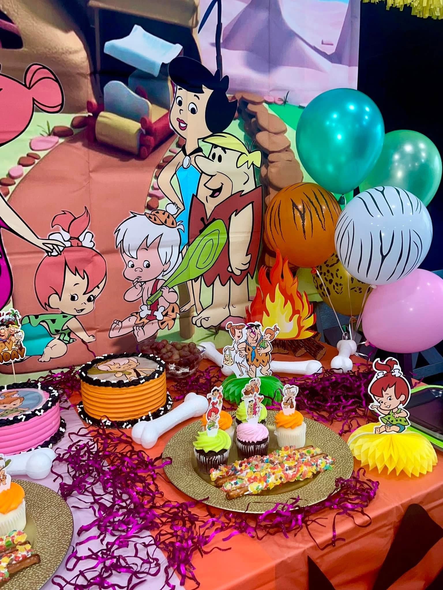 Flintstones themed birthday party table with cupcakes, decorated cakes, and character cutouts.